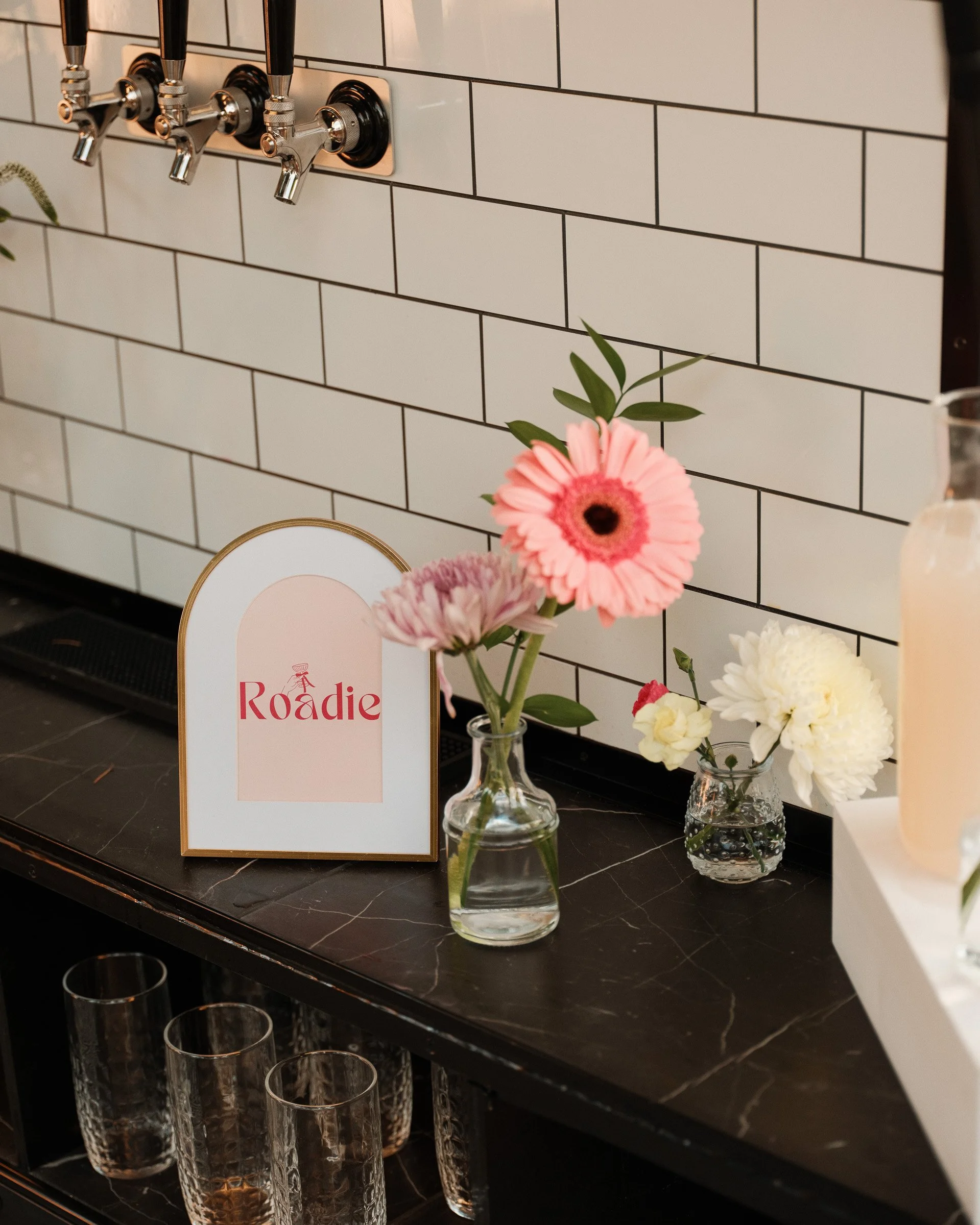 Three beer taps mounted on a white tiled wall, with several empty patterned glasses and a small bouquet of flowers on a dark countertop.