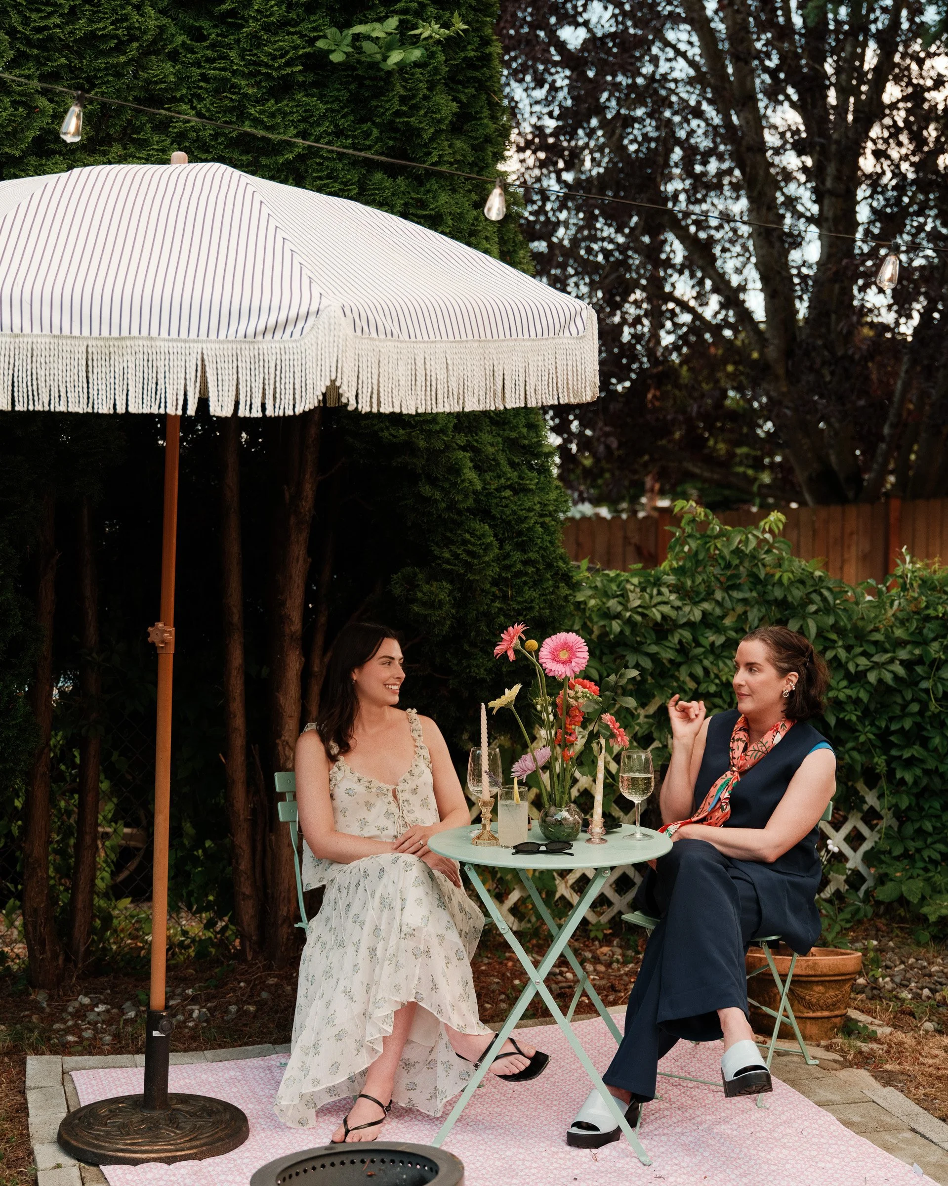 Two women sit at a small round table outdoors under a white umbrella with fringe. The woman on the left wears a floral dress and the woman on the right wears a dark sleeveless top with a colorful scarf, gesturing with her hands. The table holds flowe