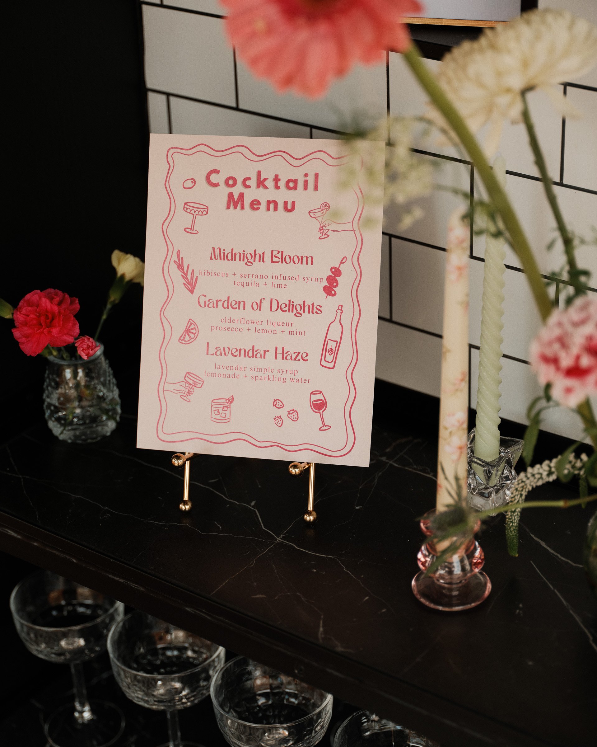 A cocktail menu featuring drinks called Midnight Bloom, Garden of Delights, and Lavendar Haze, displayed on a stand with flowers and glasses nearby.