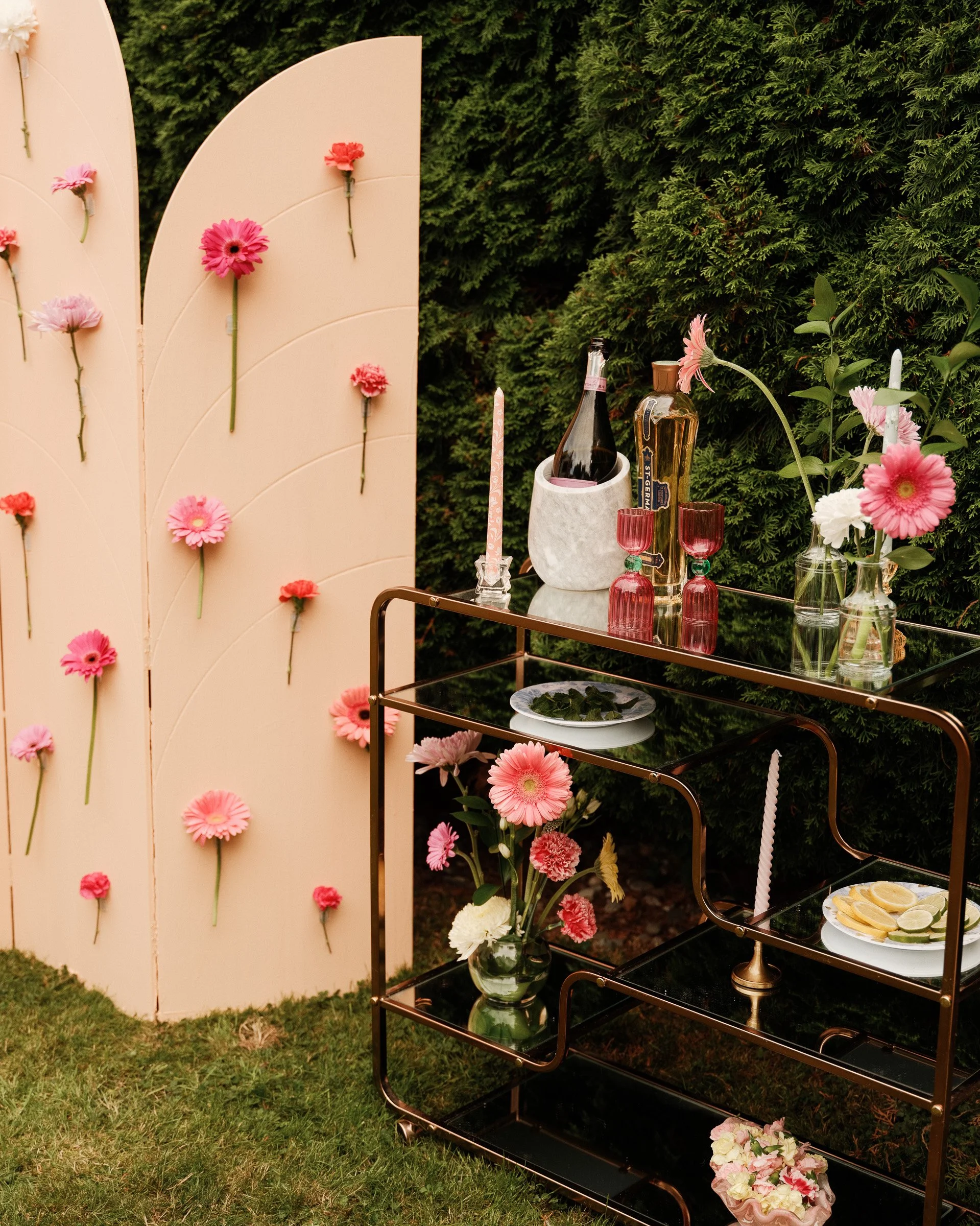 A garden decorated for a celebration features a cream-colored flower wall with pink and red carnations attached, a black metal and glass rolling bar cart with pink glasses, bottles, and flowers, and a white circular plate of lemon slices on the lower