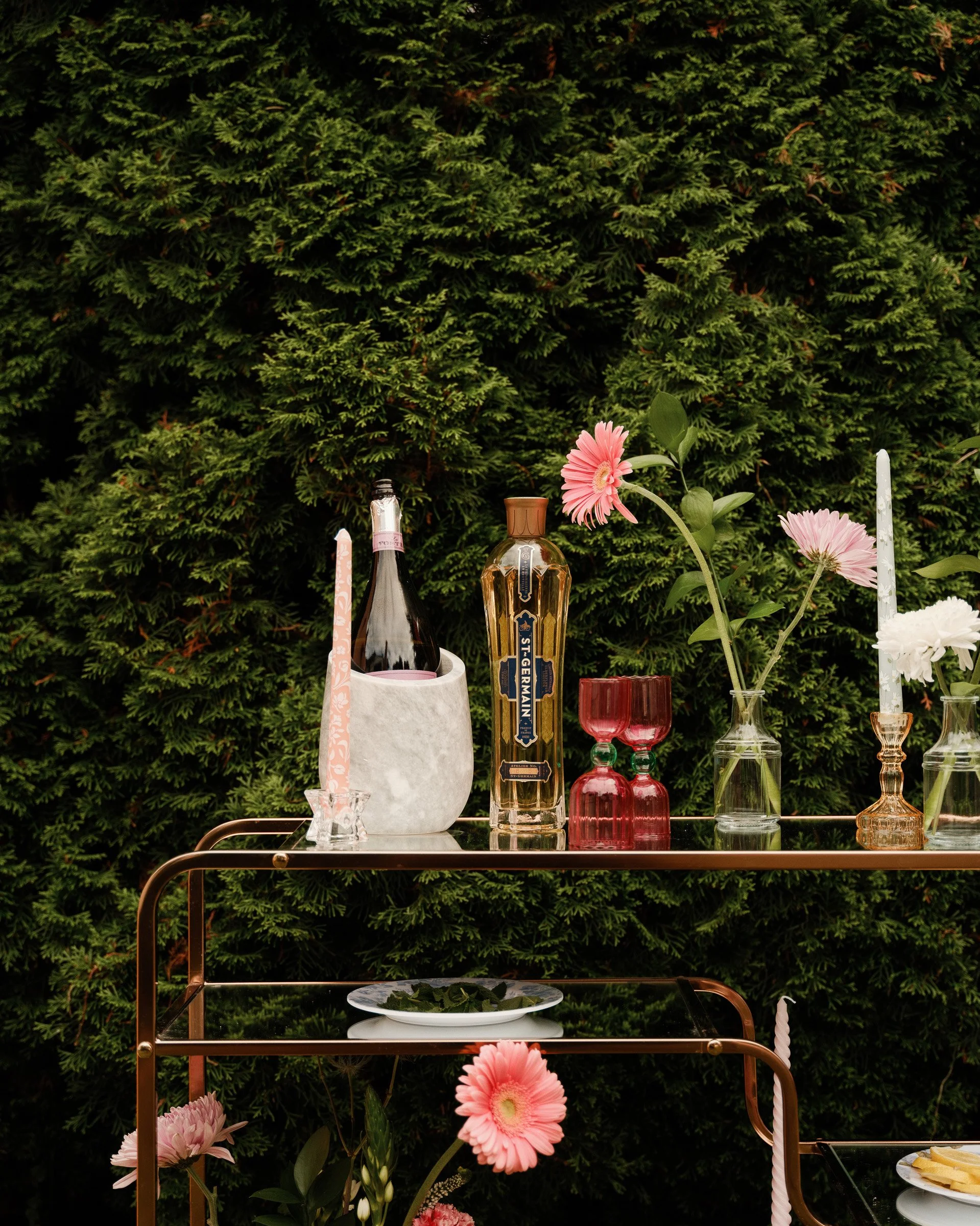 Decorative table setting outdoors with pink and white flowers, glassware, candles, and bottles against a lush green hedge background.