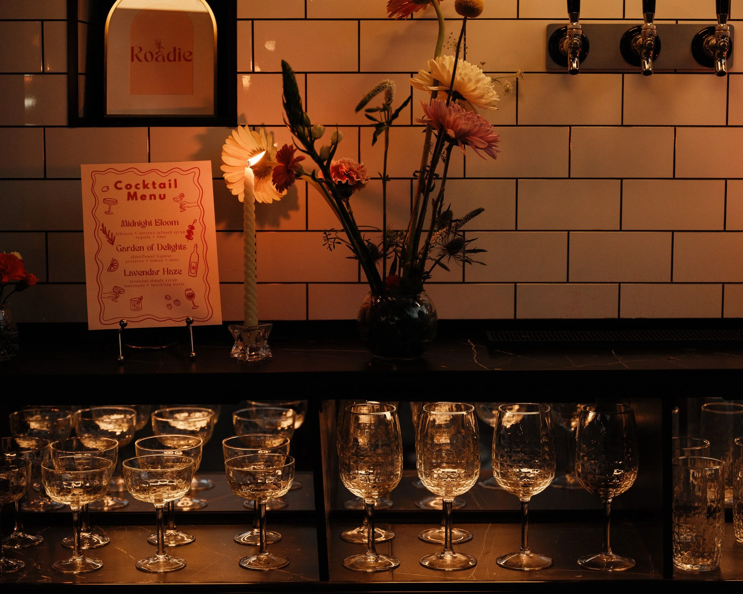 A display of empty glasses arranged on a dark surface, with a vase of flowers and a candle in the background. The wall behind has white subway tiles and there is a cocktail menu sign with a candle beside it.