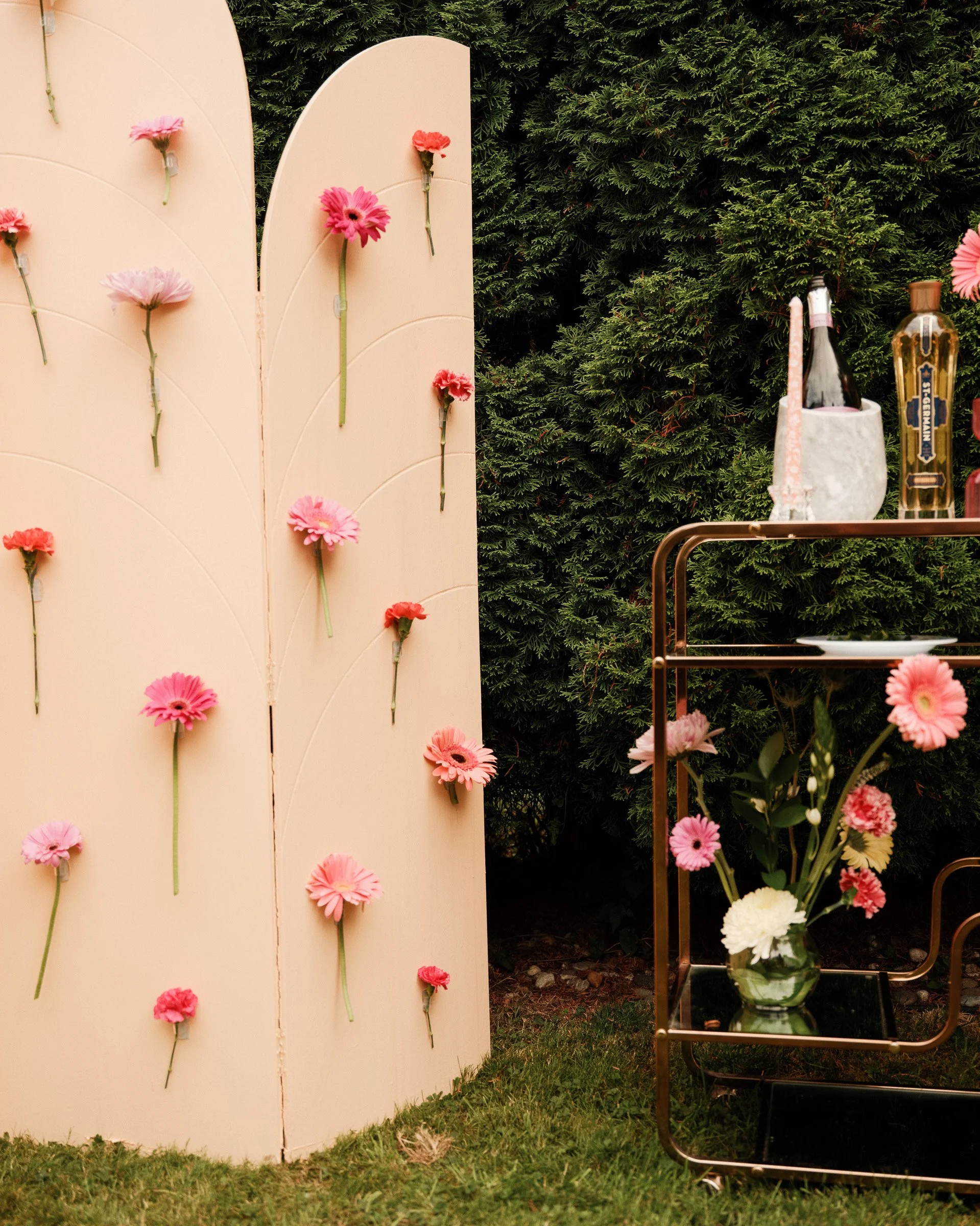 A beige cardboard display with pink and red flowers attached to its surface, set outdoors beside green bushes. Next to it, a small metal cart holds a glass vase with pink and white flowers and two bottles of alcohol, one of which is St-Germain elderf