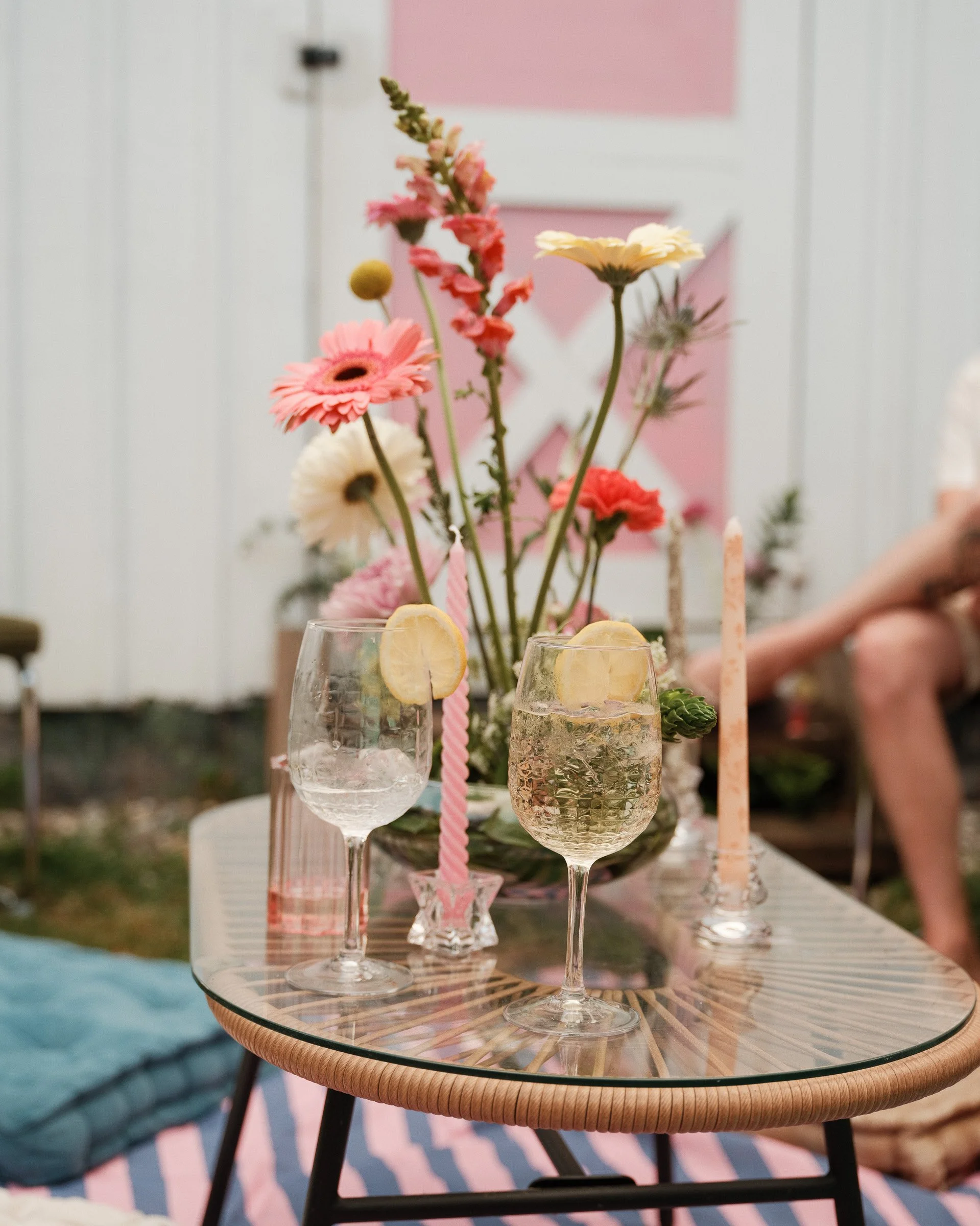 A glass outdoor table with flower centerpieces and drinks, set for a gathering, with a pink and white background.