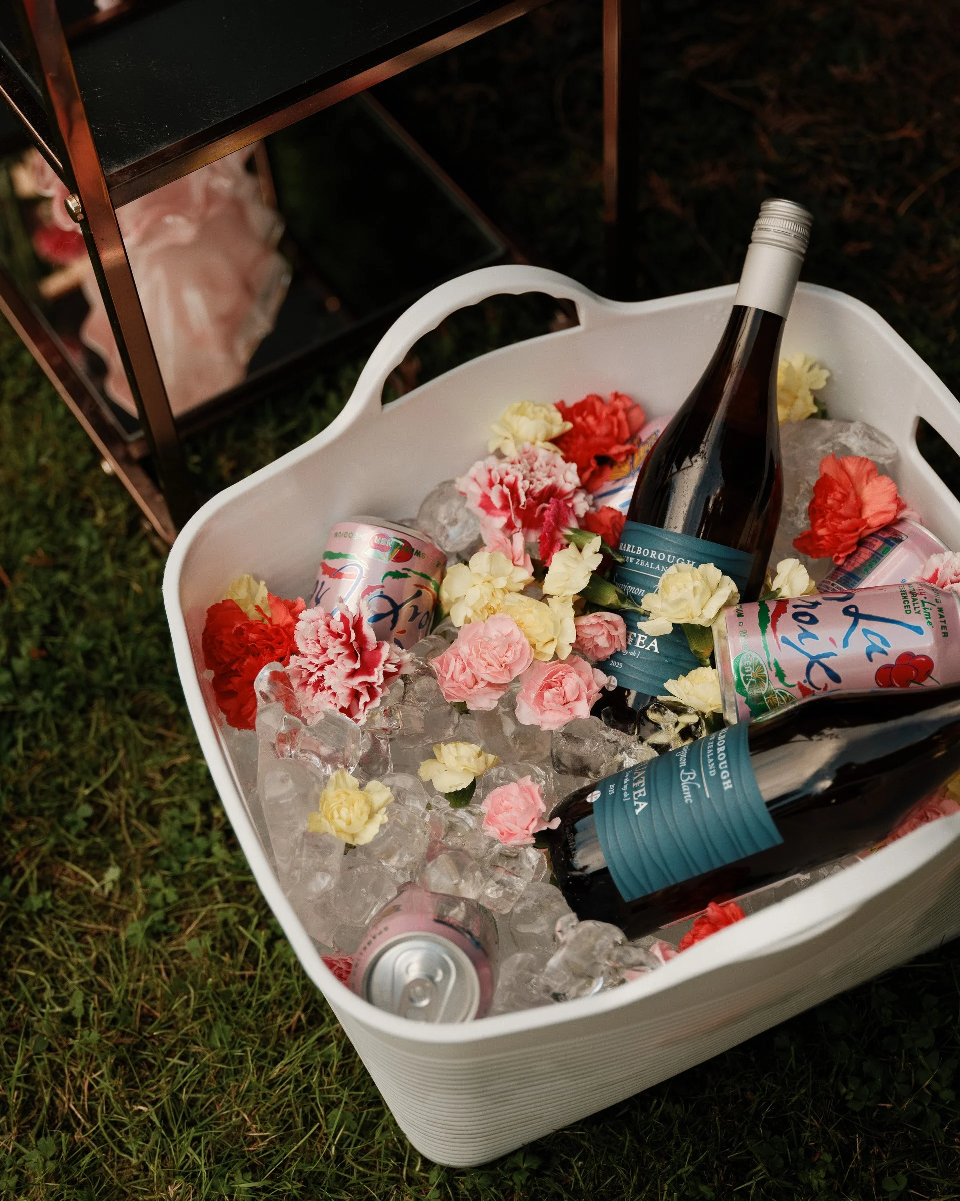 A white cooler filled with ice, featuring a bottle of Marlborough Sauvignon Blanc wine, several cans of Top Pop sparkling water, and colorful flowers, outside on grass.