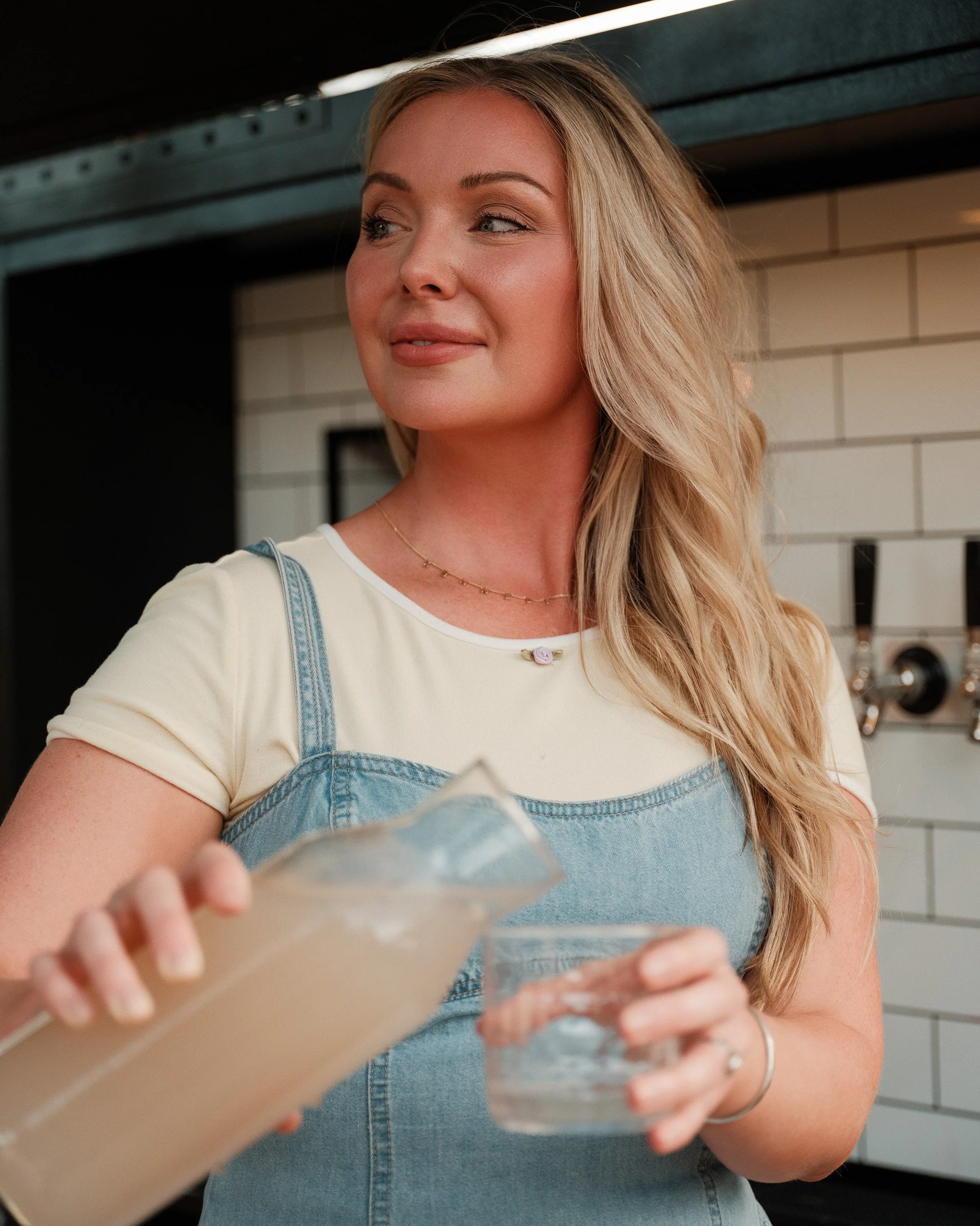 A woman with long blonde hair, wearing a white shirt and denim overalls, holding a glass of water in front of her. She is standing in a modern kitchen with white tile backsplash.