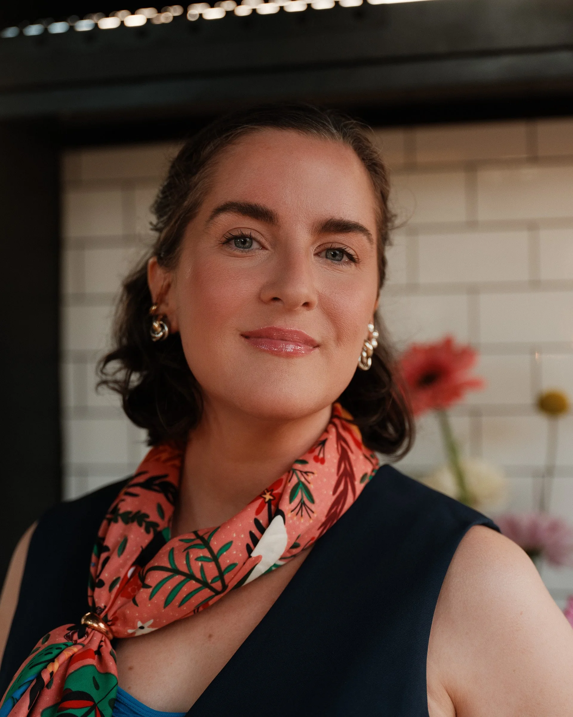 A woman with styled dark hair, wearing large earrings and a red floral scarf, smiling, in front of a white tiled wall with a red flower in the background.