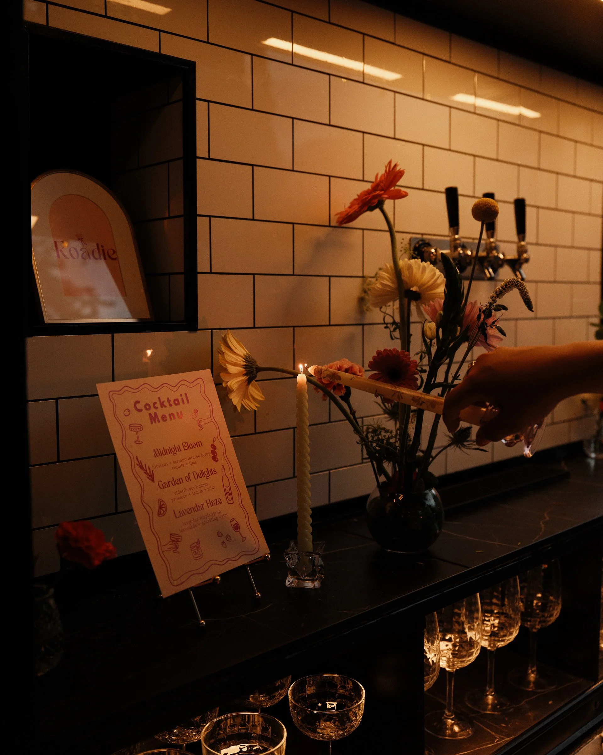 Display of cocktails, flowers in a vase, and cocktail menu on a bar counter with a tiled wall in the background.