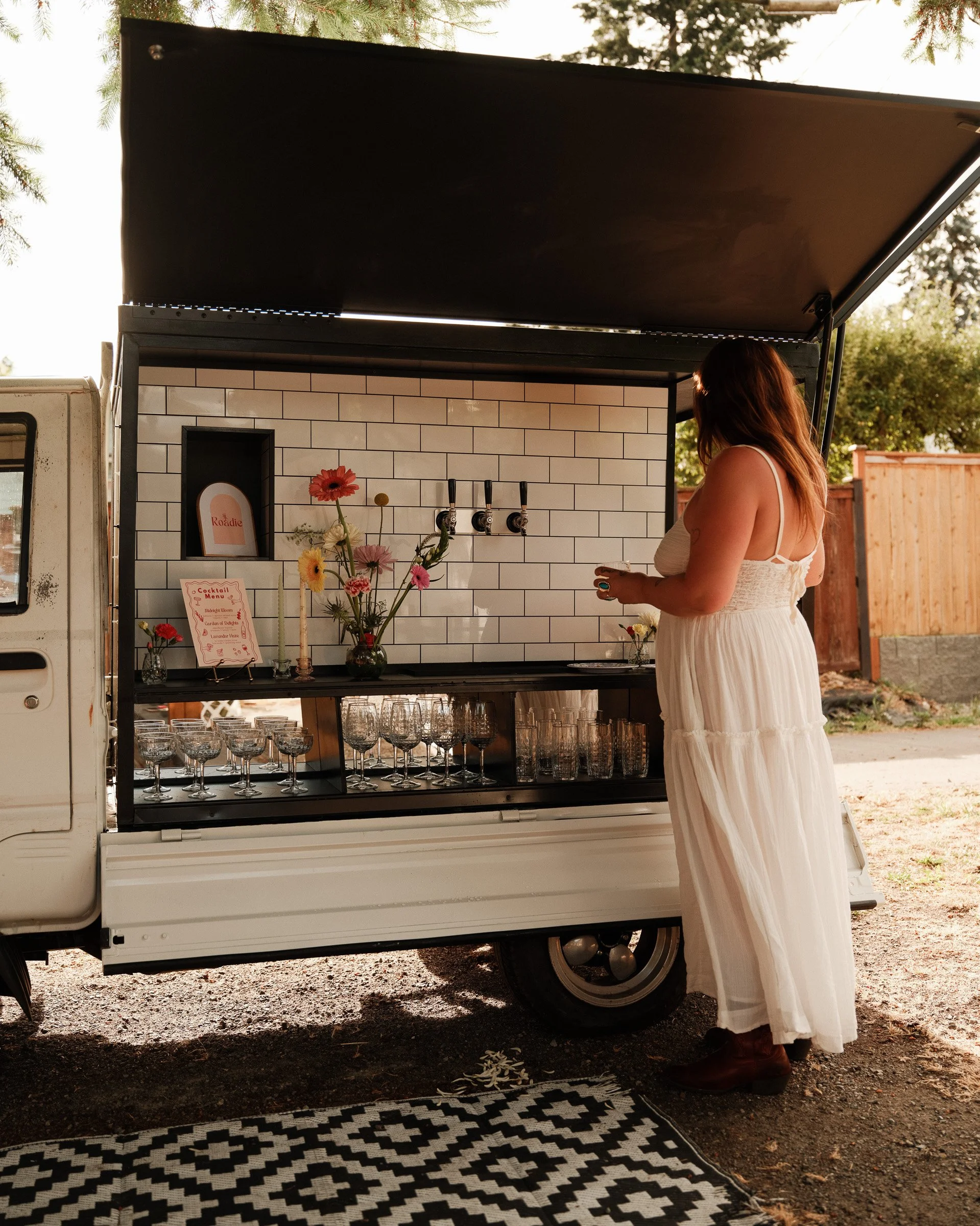 A woman in a white dress and brown boots stands at a mobile outdoor bar with a white tile backsplash, flowers, and beverage taps, under a canopy on a dirt ground. There are trees and a wooden fence in the background.