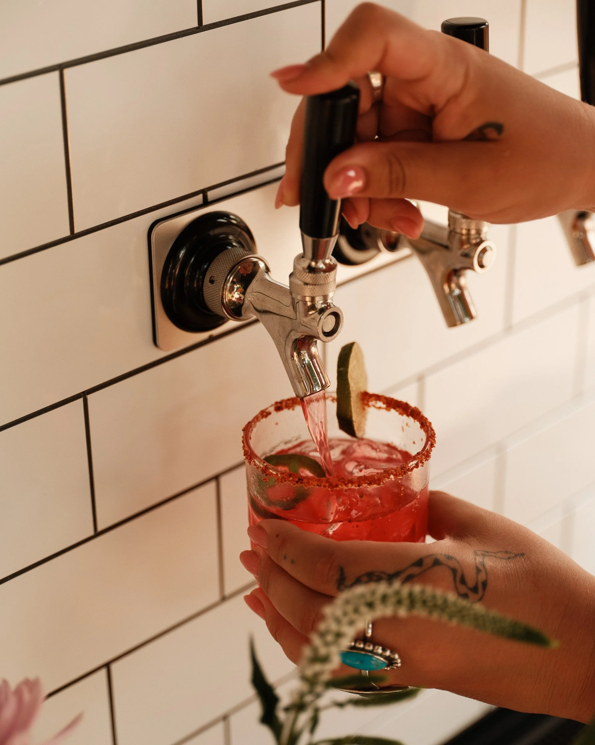 Person holding a drink with a straw, garnished with a lime and salt rim, as a tap pours pink liquid into the glass, against a tiled wall background.