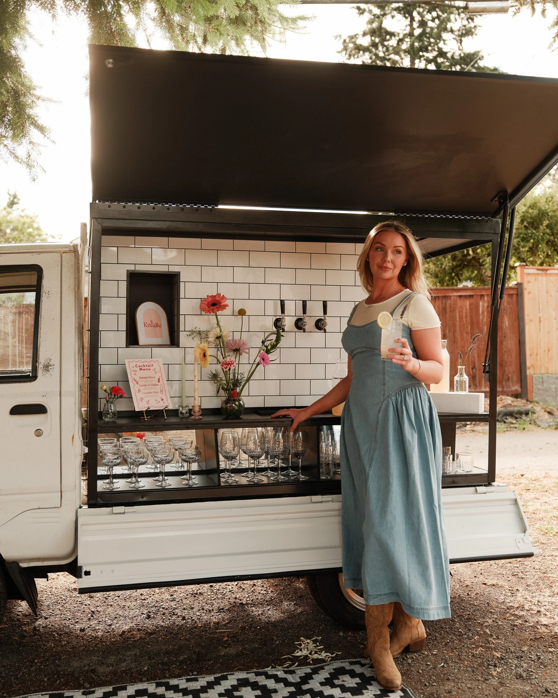 A woman in a denim dress and cowboy boots stands beside a mobile bar in front of a food truck, holding a drink with lemon slice, outdoors with trees and a wooden fence in the background.