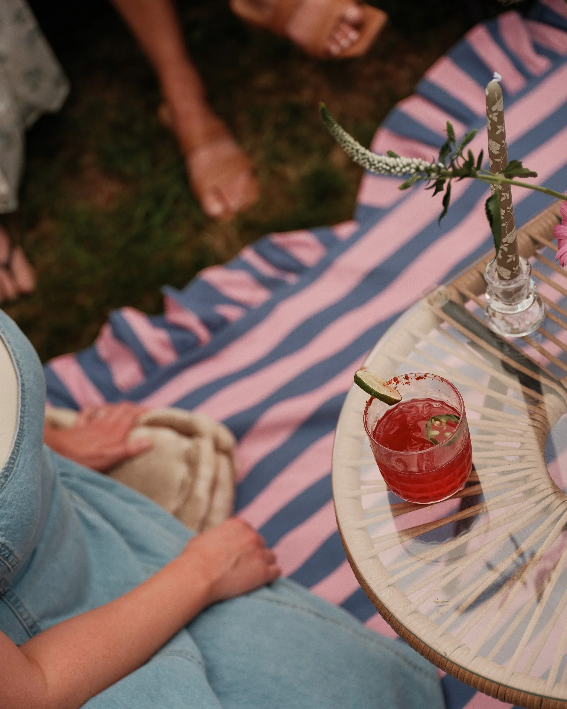 A person sitting on a pink and blue striped blanket, holding a glass of red cocktail with lime and cherry garnishes on a small wooden table, with a floral candle holder nearby.