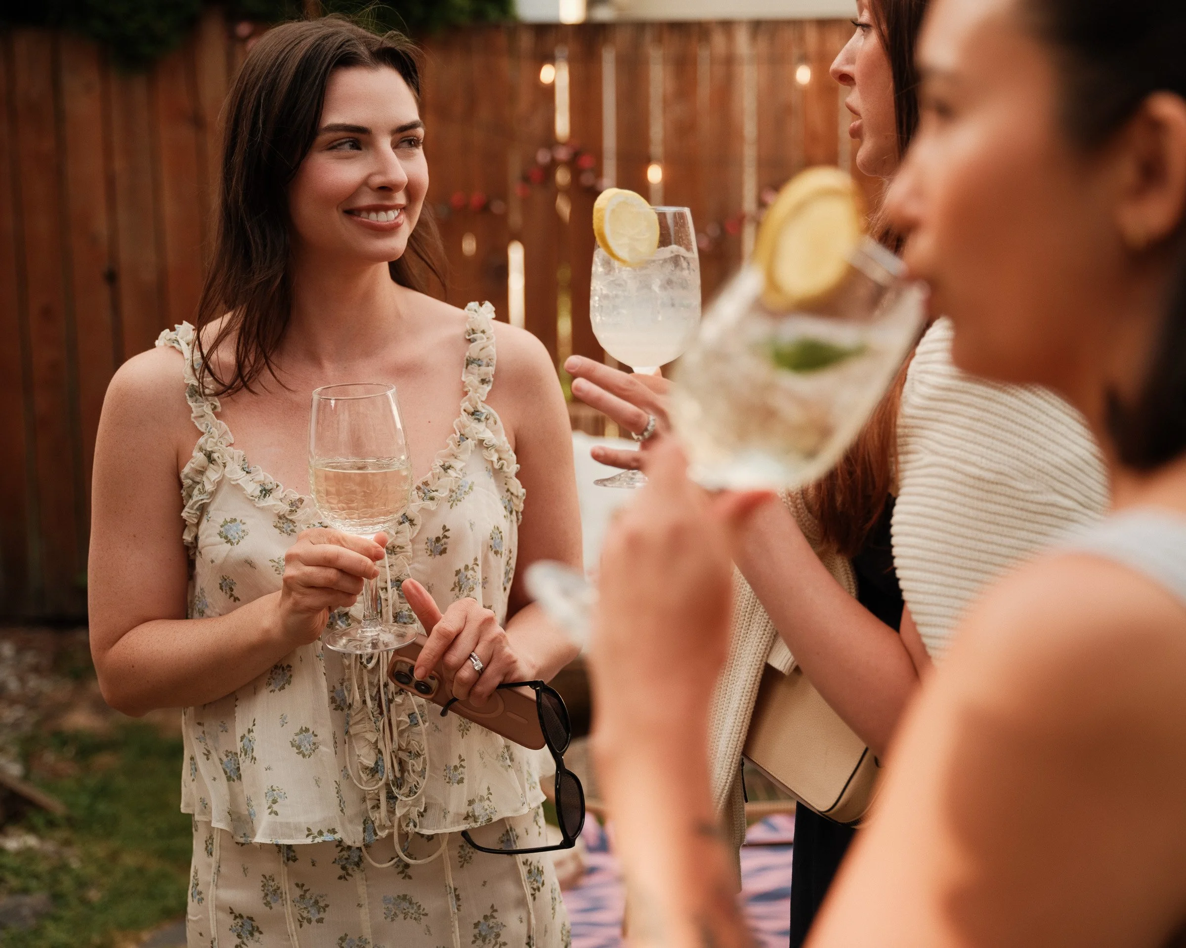 Women at an outdoor gathering holding cocktails with lemon slices, with a wooden fence and string lights in the background.