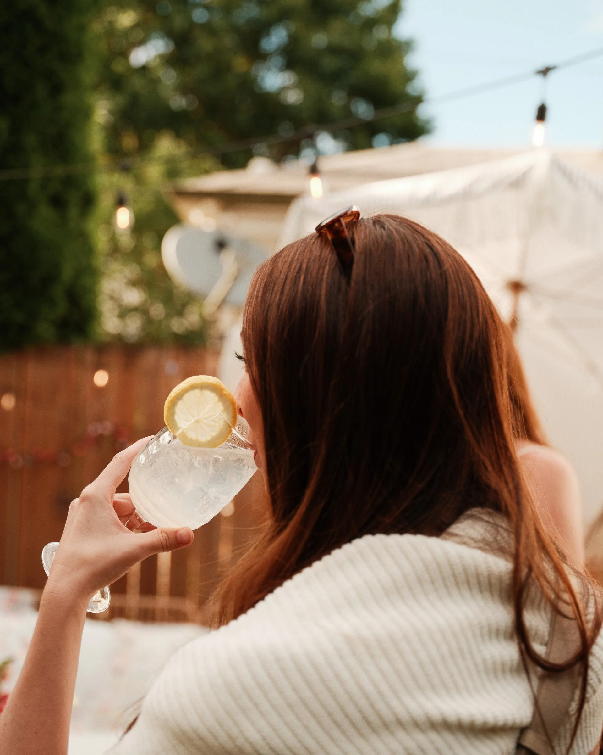 A woman with long reddish-brown hair holding a glass of lemonade with a lemon slice garnishing the rim, outdoors with blurred background of trees, string lights, and a white canopy.