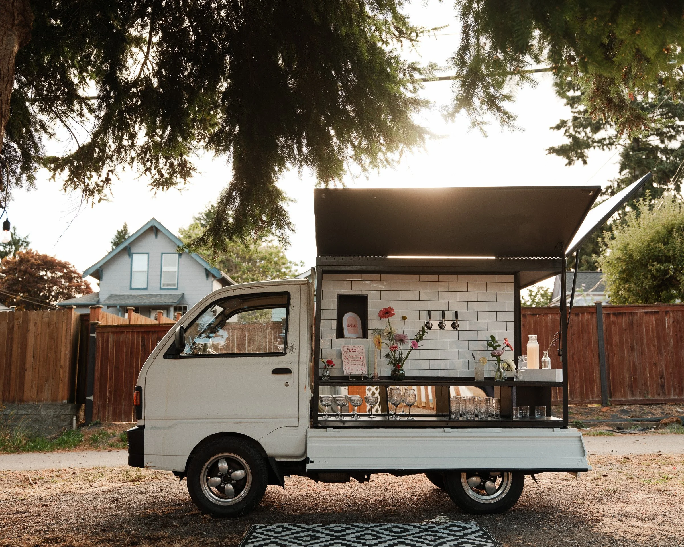 A white food truck with a black serve window parked in a residential neighborhood with trees and houses in the background. The truck has a floral display with glasses and bottles on the side and a checkered black-and-white rug in front.