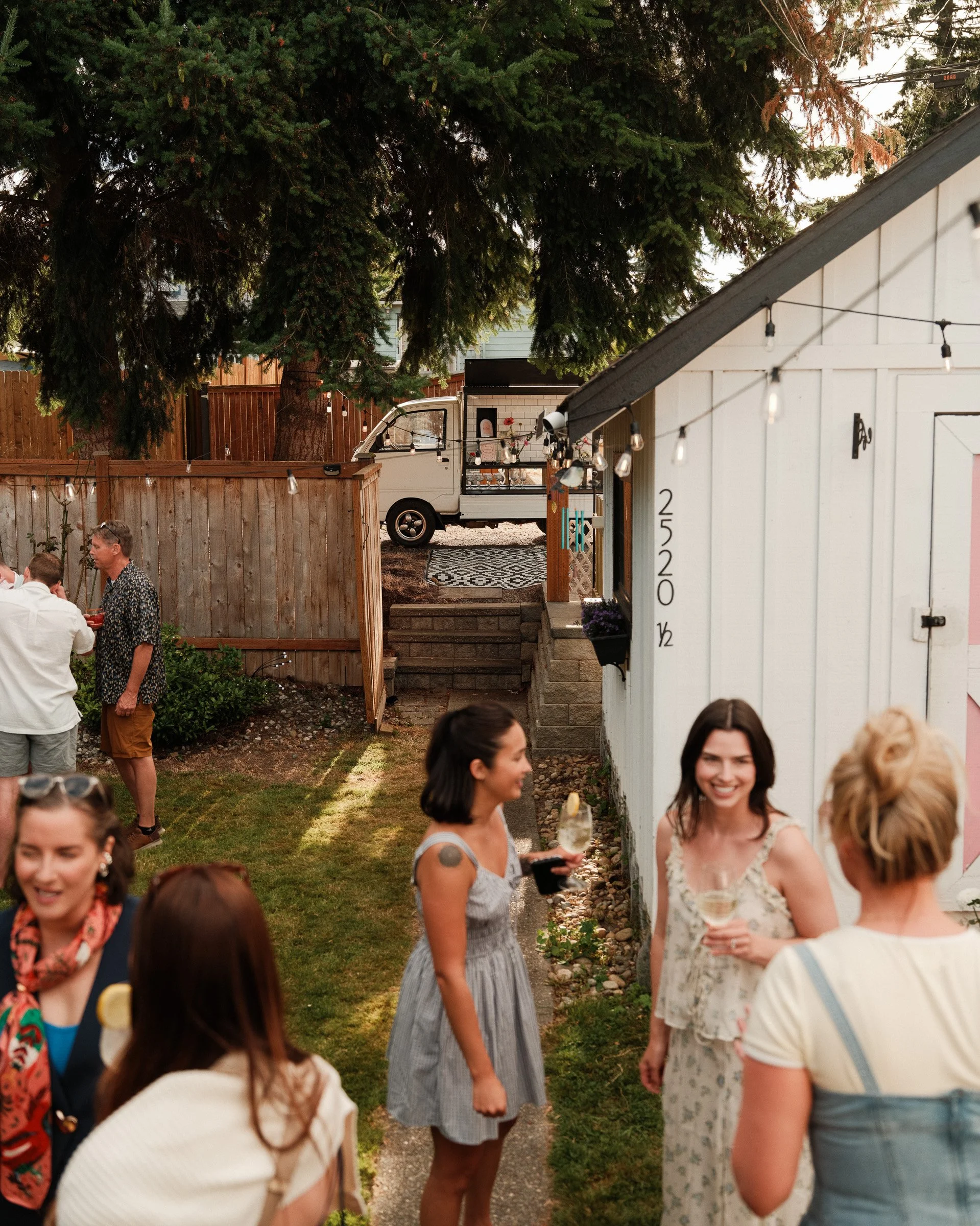 People socializing in a backyard during a gathering with string lights, a food truck, and a large tree.