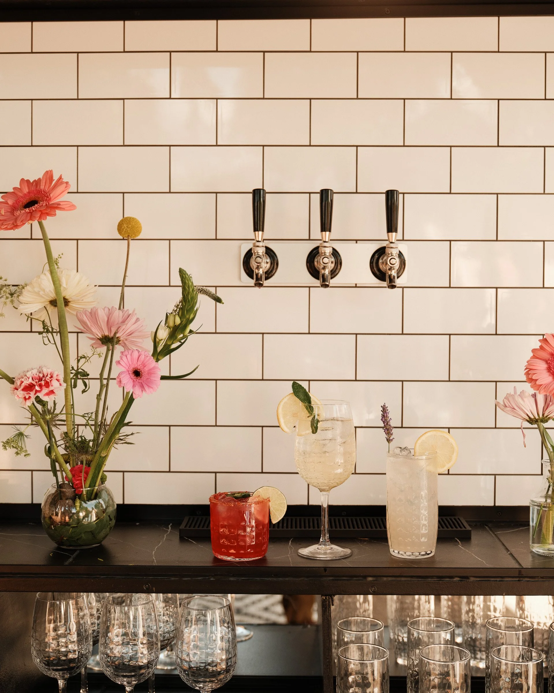 A bar setup with a white tiled wall in the background, decorated with a glass vase filled with pink and white flowers on the left side, and a variety of beverages in glasses with lemon slices on the right side. There are three beer taps mounted on th