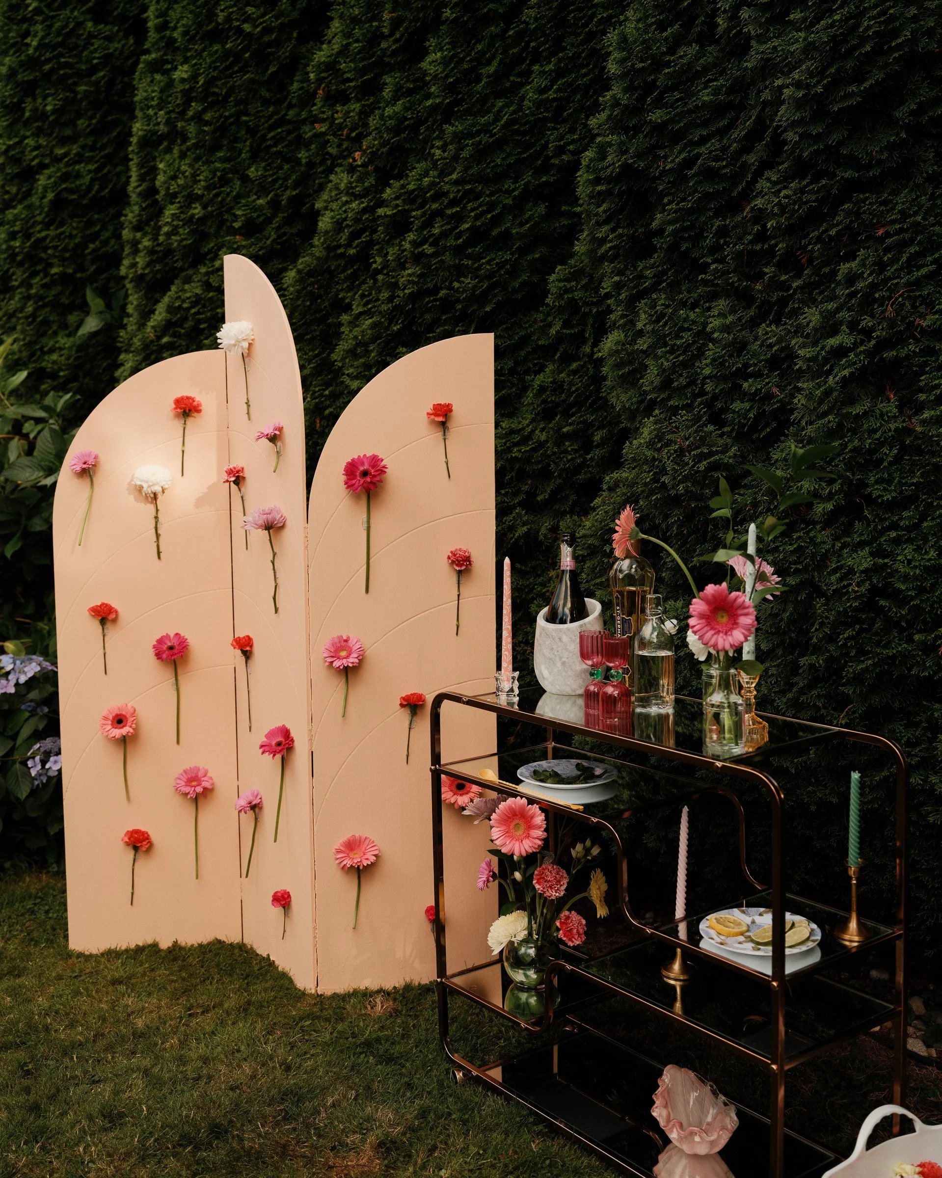 Outdoor party setup with a pink flower display and a black metal serving cart holding drinks, candles, and plates, against a backdrop of lush green bushes.
