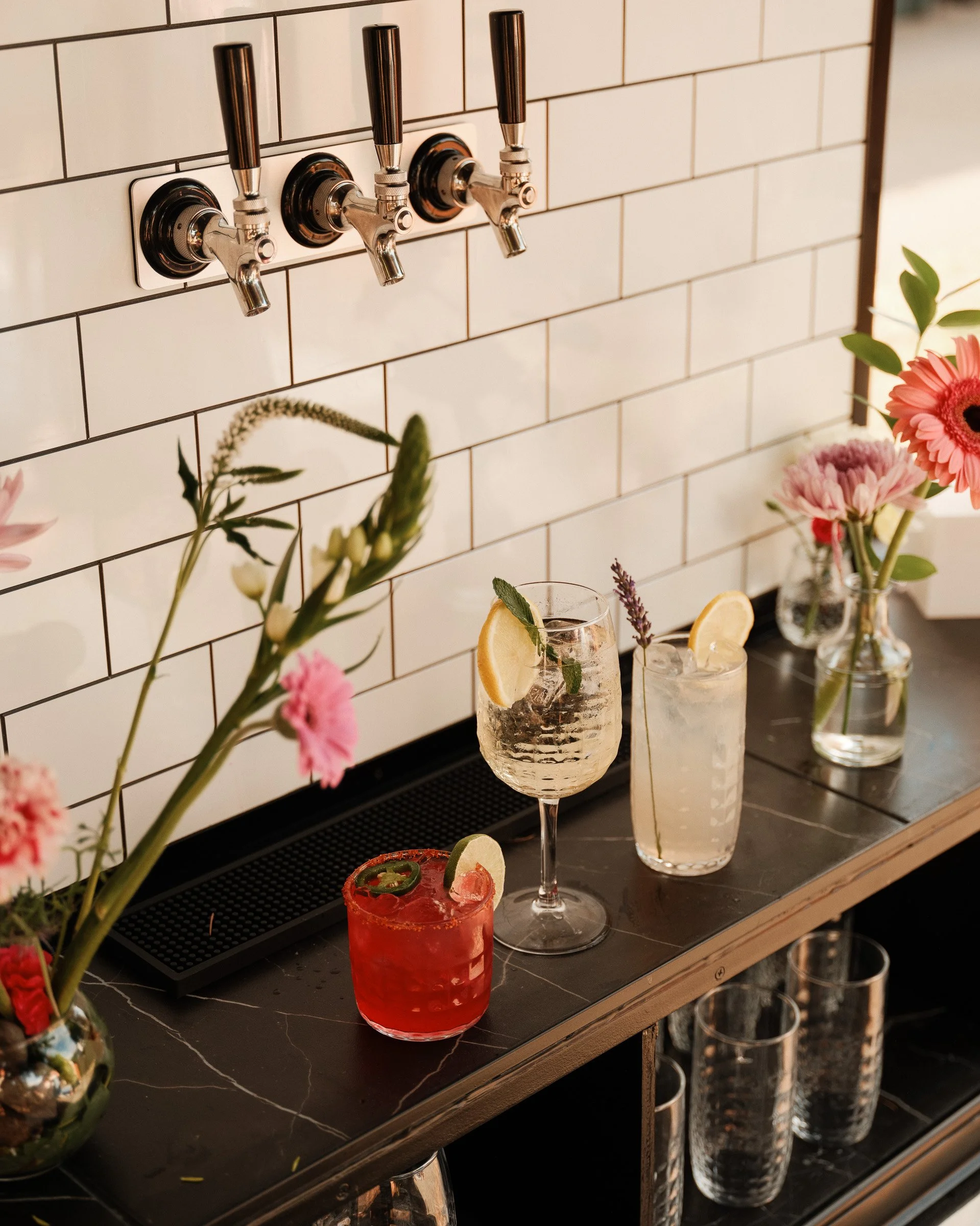 Close-up of bar counter with three cocktail glasses and fresh flowers, with white tiled wall and beer taps in the background.