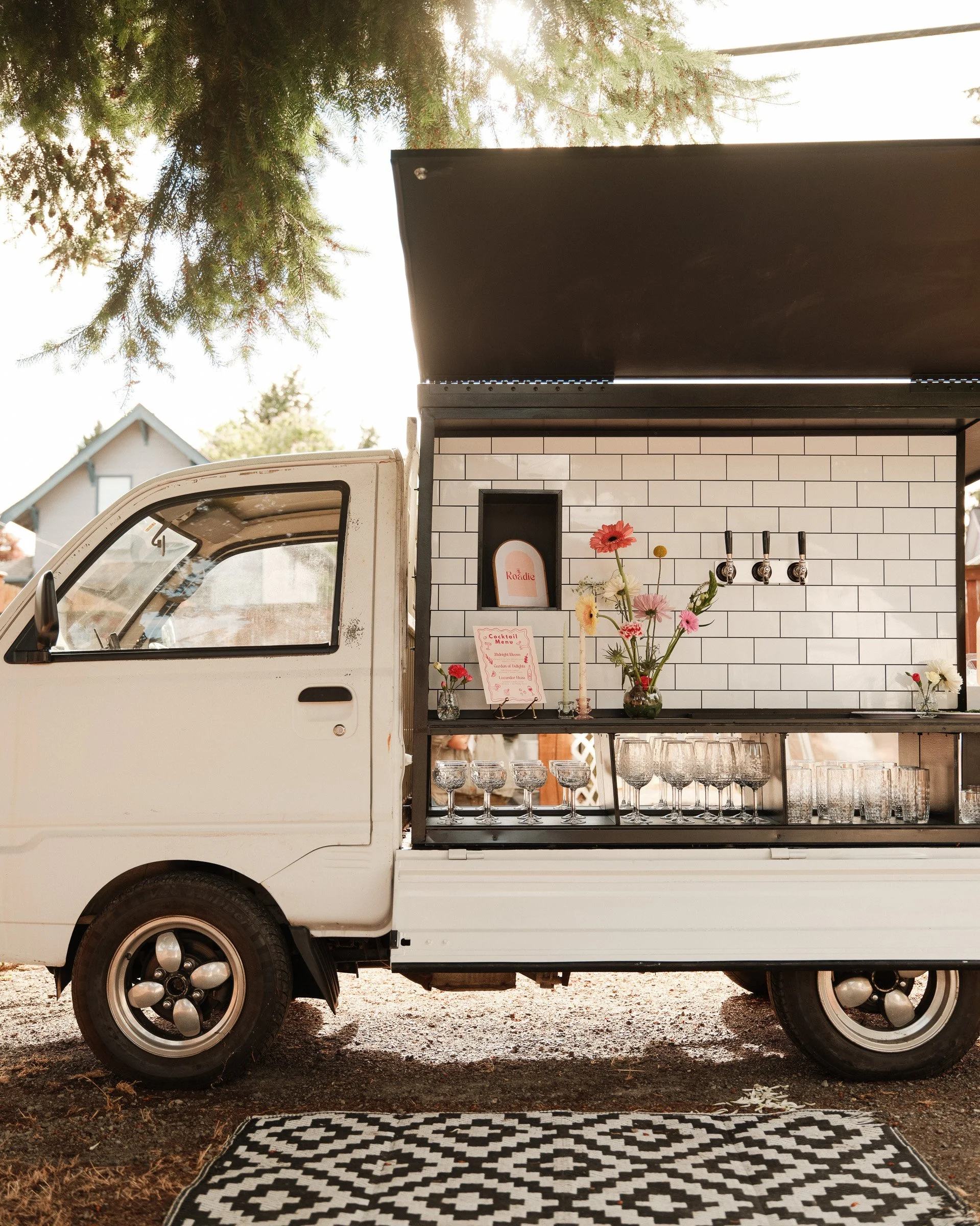 A food truck with a white brick exterior serving drinks, decorated with flowers, set outside with trees and a house in the background, and a black and white patterned rug in the foreground.