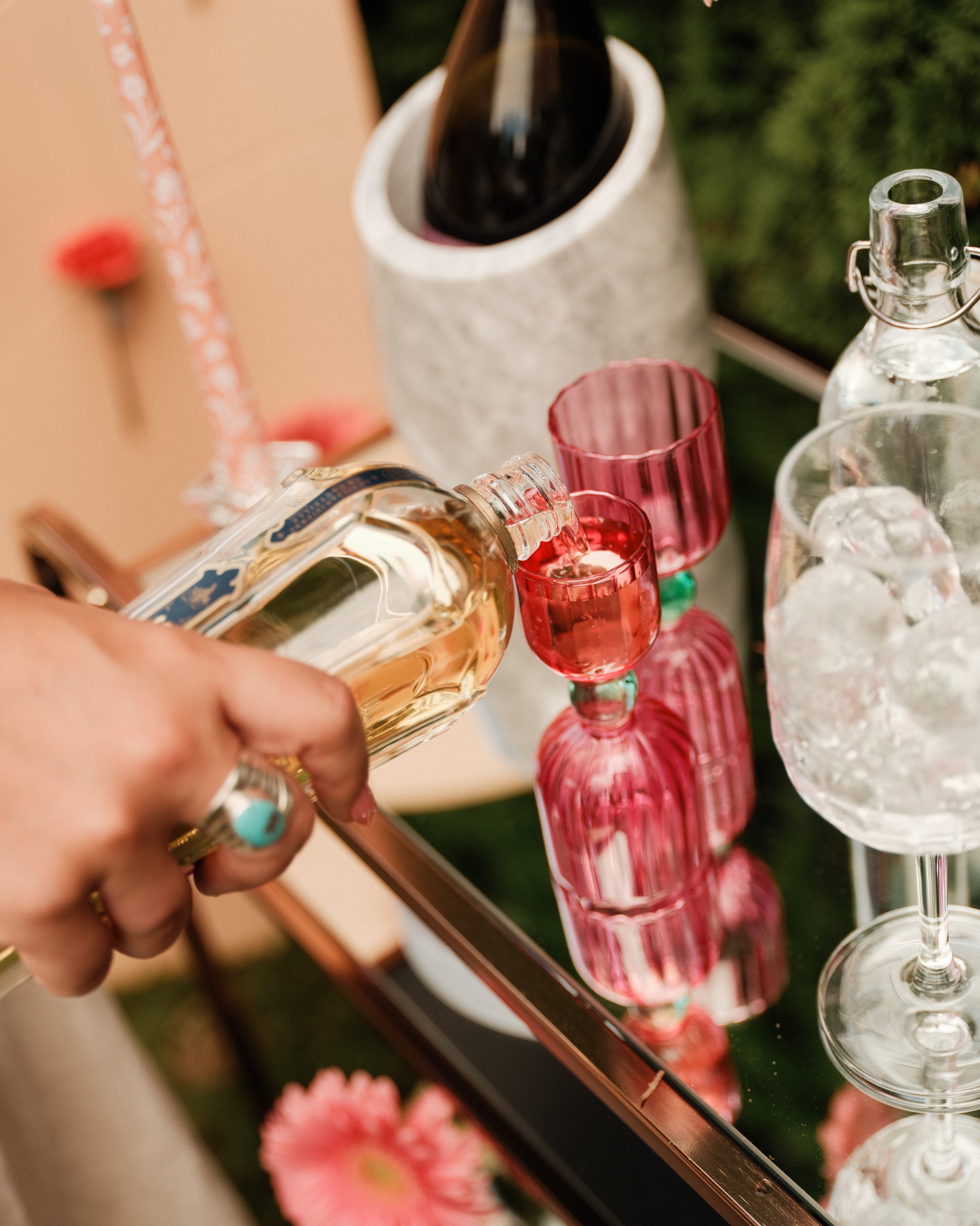 Close-up of a person pouring a clear beverage from a glass bottle into a pink glass at a party. Several other pink glasses and bottles are on the table, with a blurred background of greenery and pink flowers.
