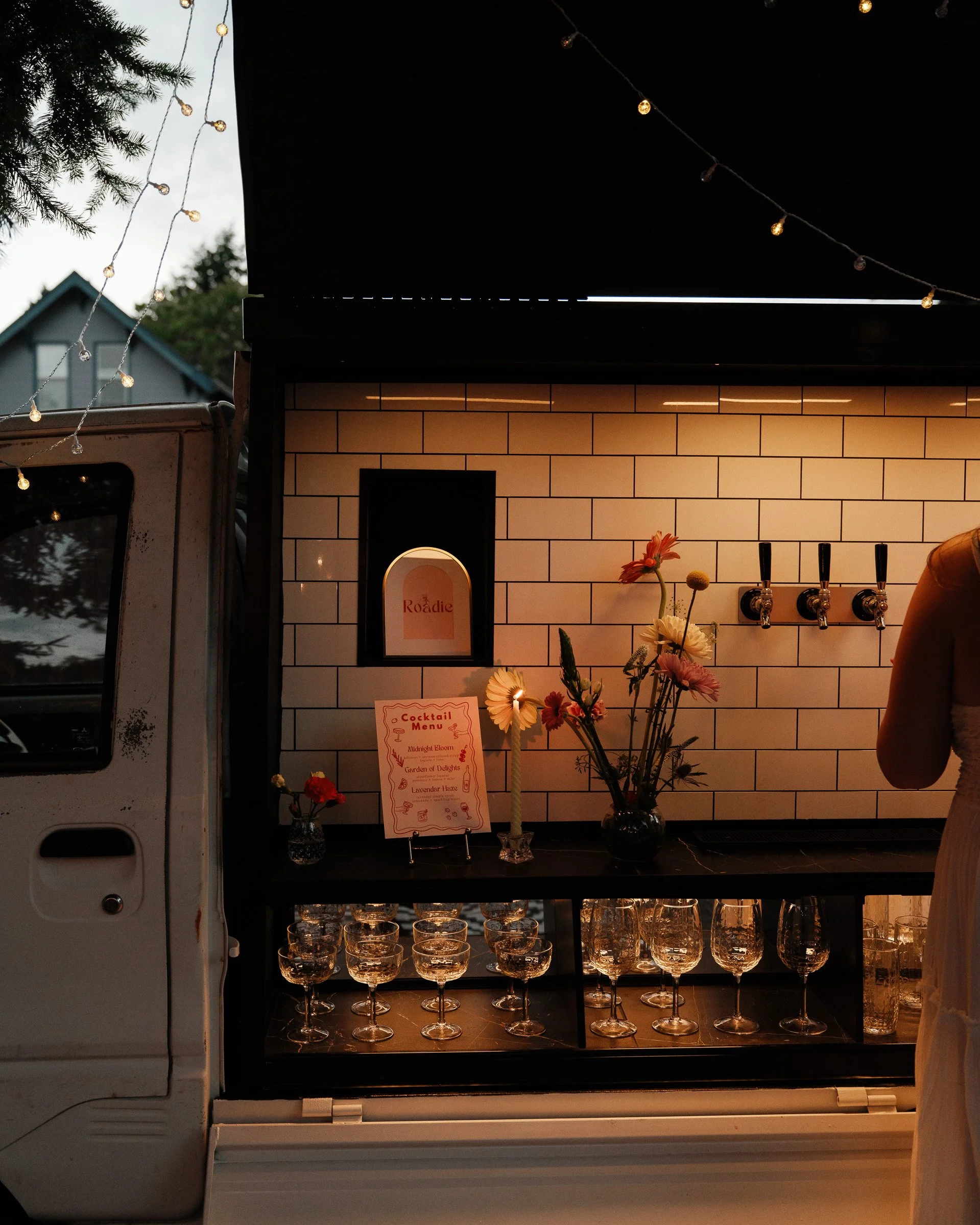Bar area with a black counter displaying wine glasses and a vase with flowers, a menu sign, and a small framed poster on the wall. String lights are hanging above, and a tree and part of building are visible outside in the background.