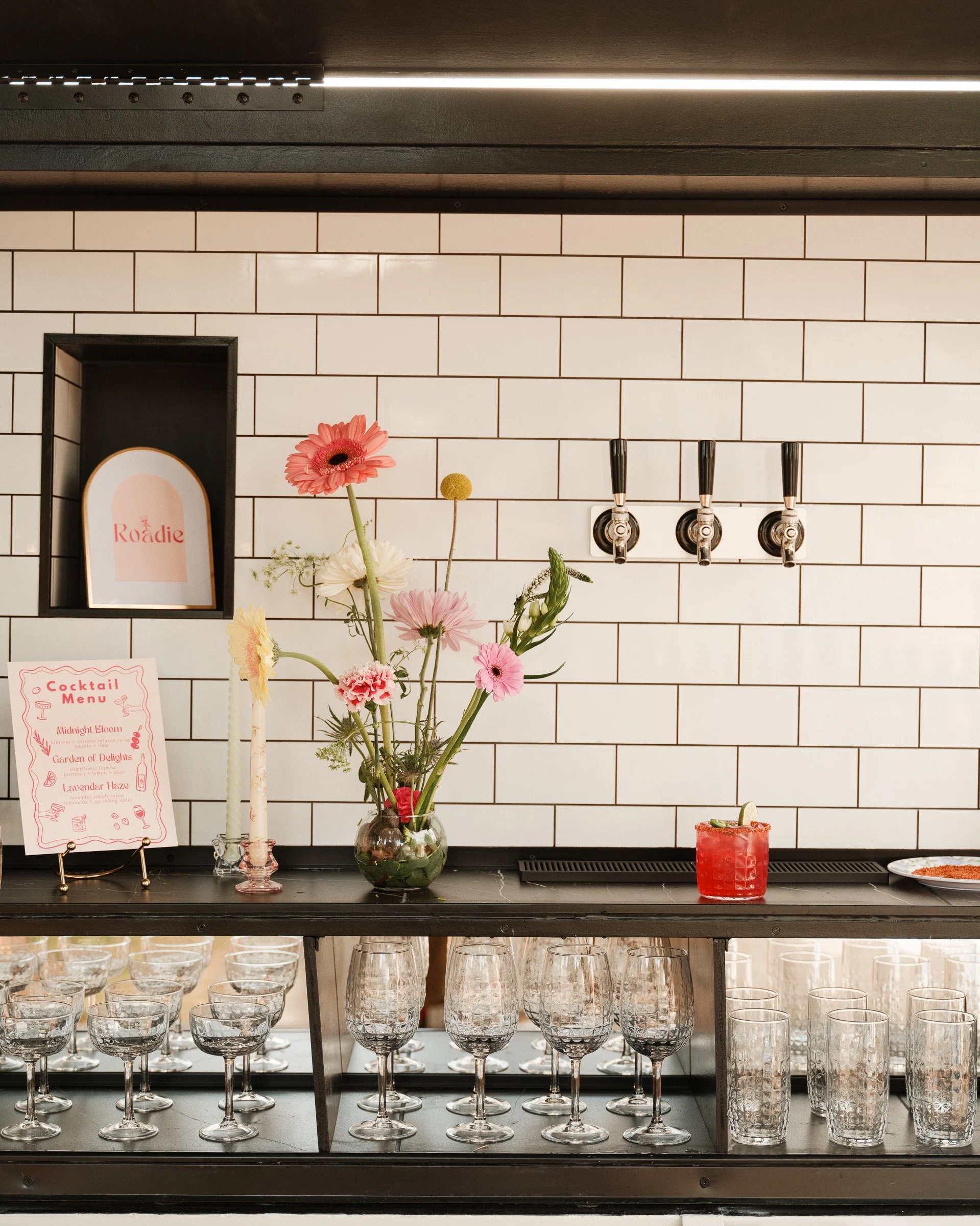 Bar setup with a bouquet of pink, white, and yellow flowers in a glass vase, cocktail menu, several wine glasses and water glasses on shelves, black tiled wall, and a red cocktail drink on the bar counter.