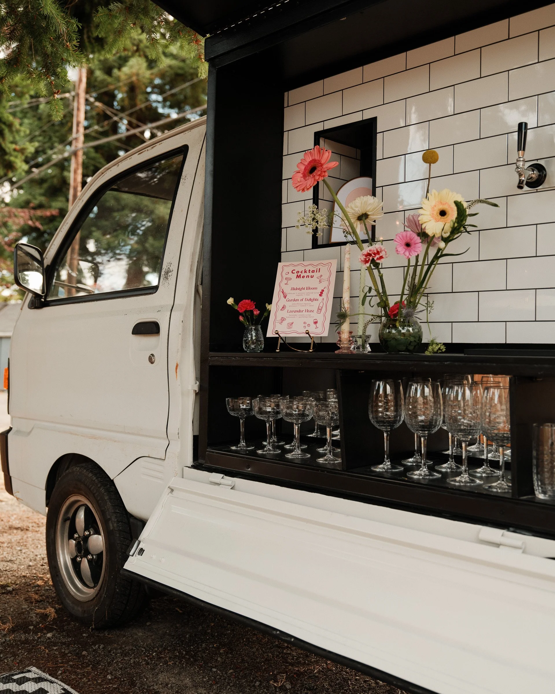 A food truck with a black interior serving area filled with various flowers in glass vases and a menu sign. The truck is white with a black trim, and there is a forest background with trees and power lines.
