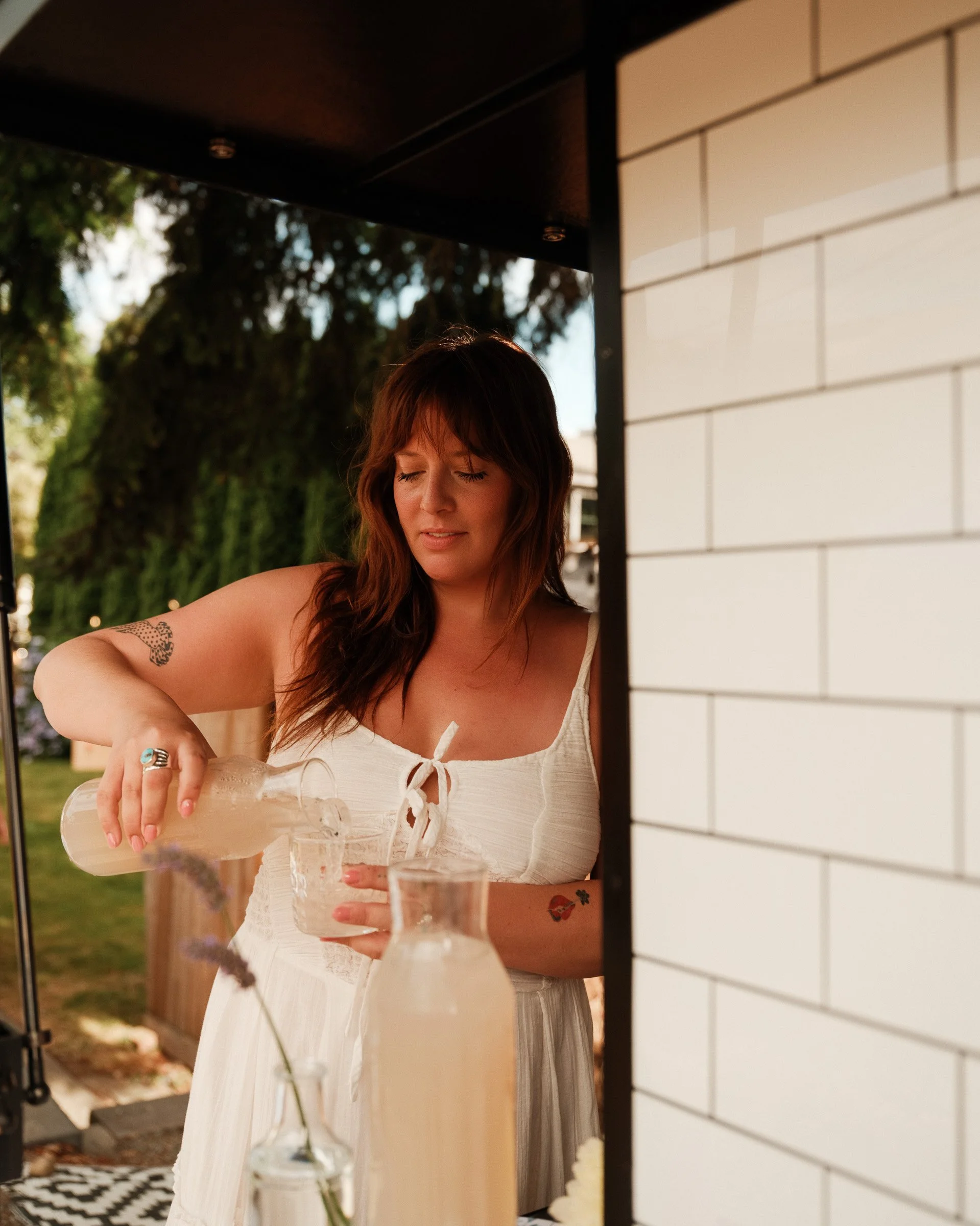 A woman with red hair and tattoos is pouring a drink into a glass at an outdoor gathering, with trees and a white brick wall in the background.