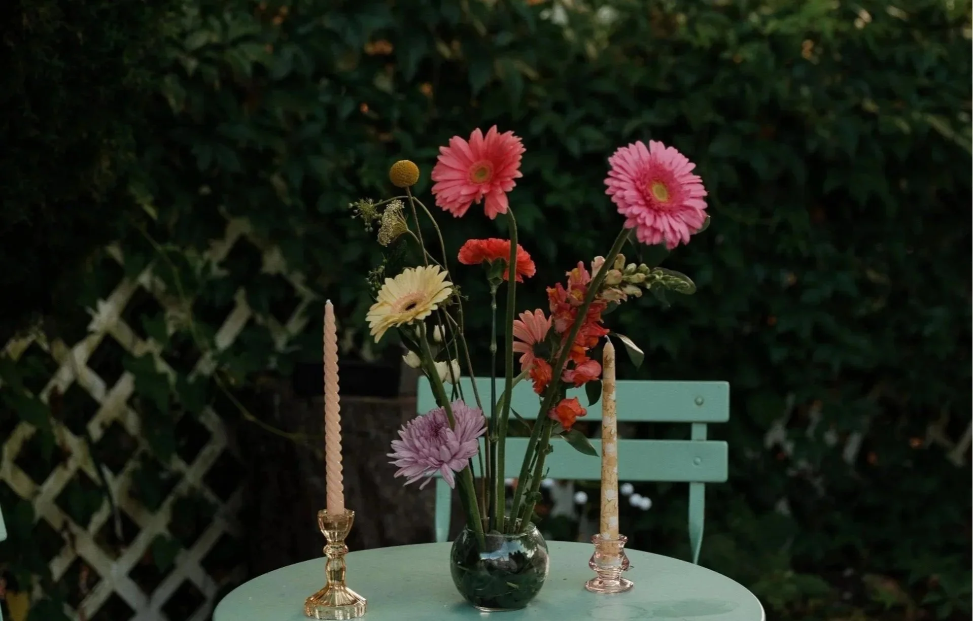 Fresh flowers in a vase on a light green outdoor table, flanked by two tall candles in decorative holders, with a green bench in the background.