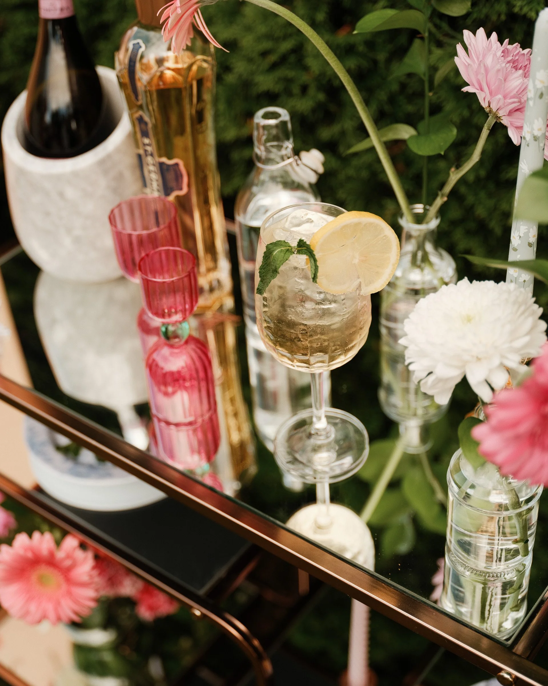 A close-up of a mirror reflecting a table of flowers, bottles, and glasses with a pink flower in a vase and a slice of lemon in a glass of water