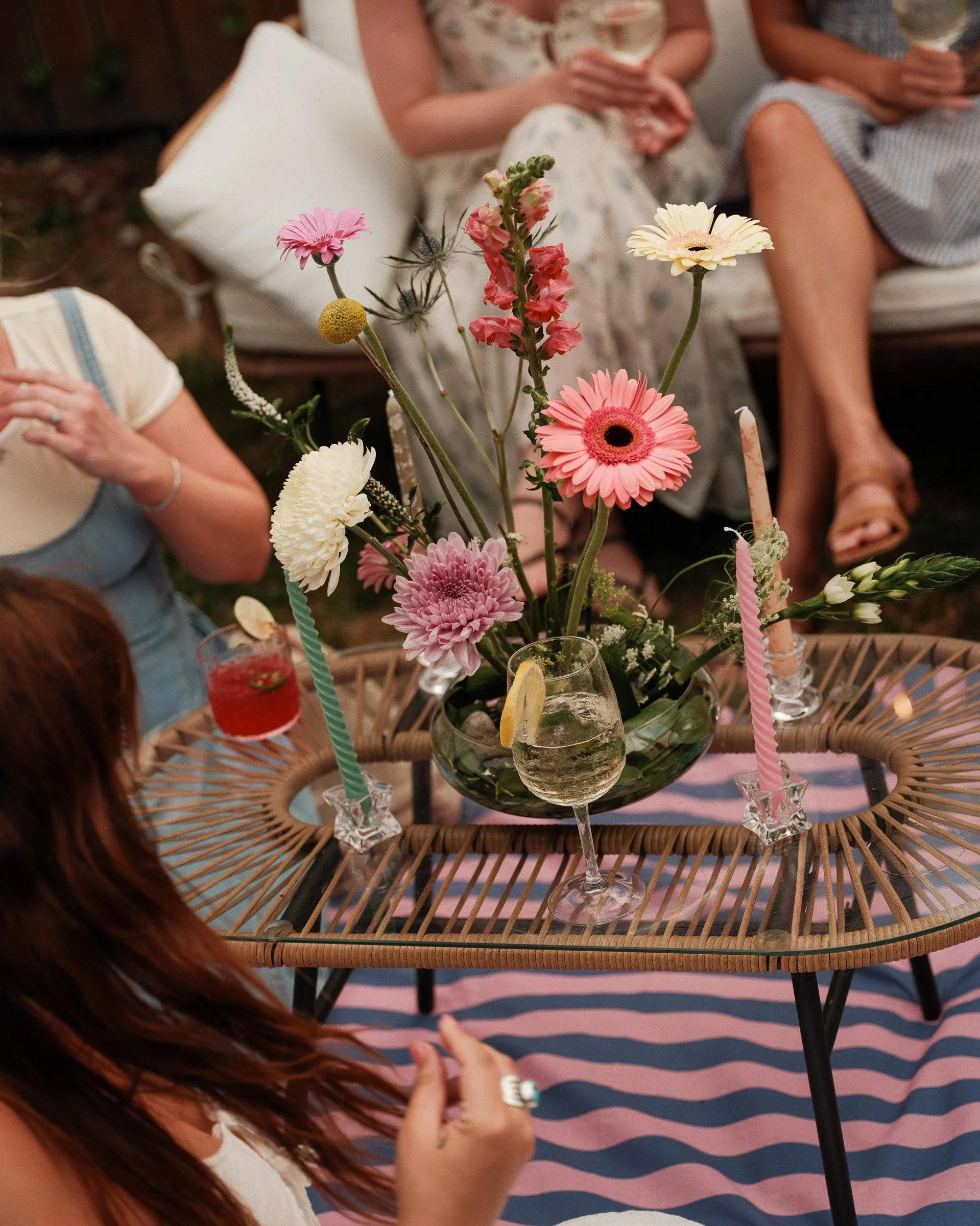 A centerpiece of pink, yellow, and white flowers on a wicker table with drinks and people sitting around, some holding glasses of wine.