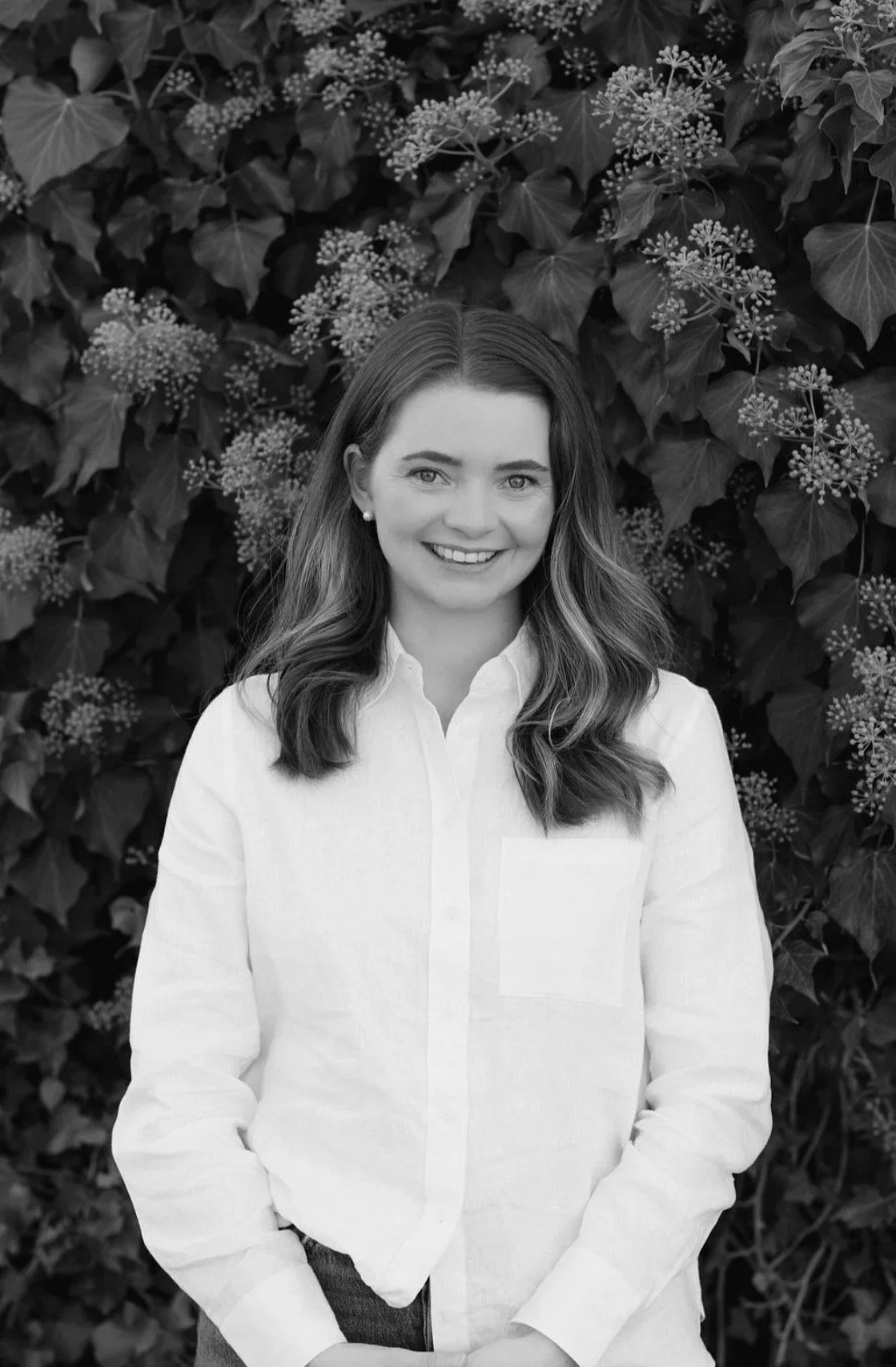 A black-and-white portrait of a woman with shoulder-length wavy hair, smiling, wearing a white button-up shirt, standing in front of leafy plants with small flowers.