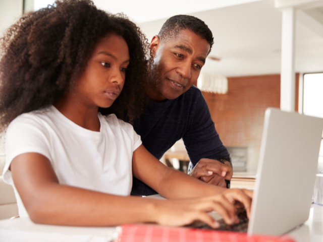 A man and young girl working together on a laptop at a table in a home setting.