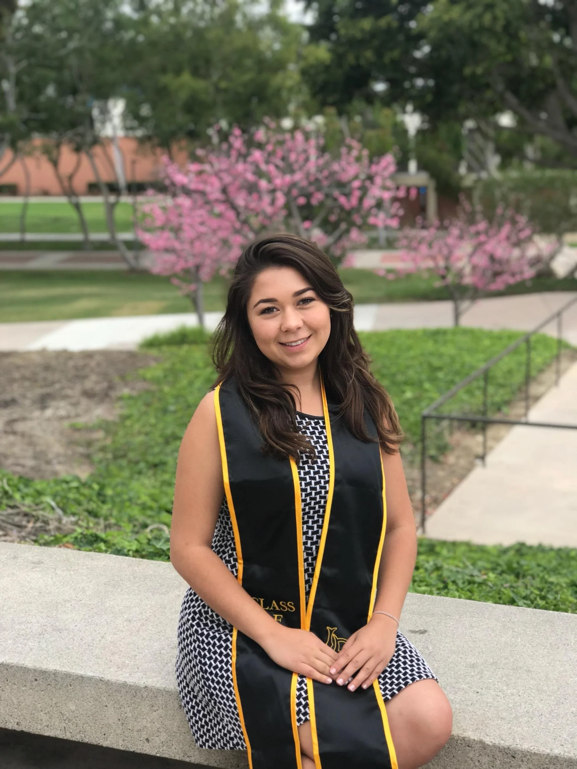 A woman in a graduation gown and cap sitting outside on a concrete bench, smiling, with pink flowering trees and green trees in the background.