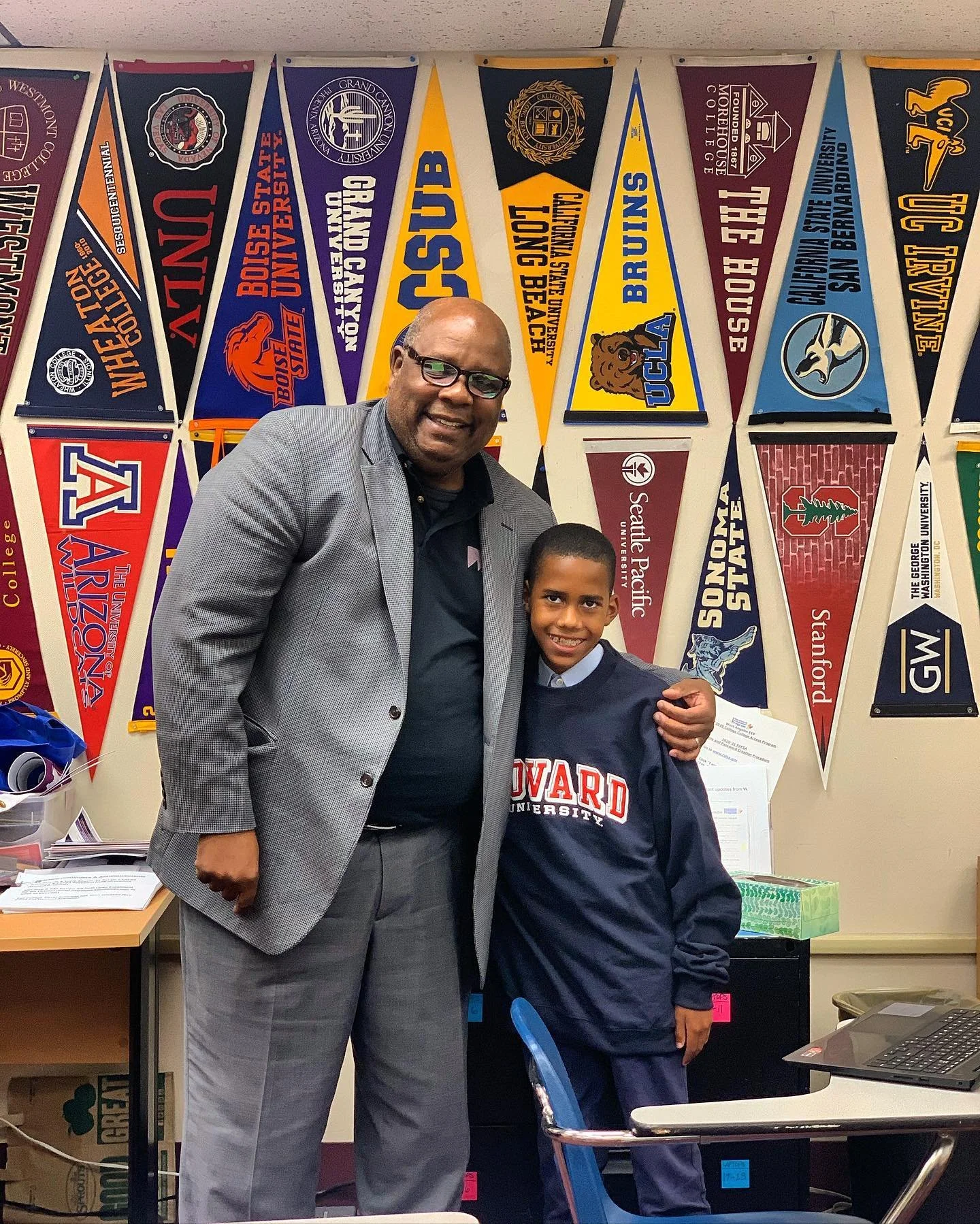 A man wearing glasses and a gray suit standing next to a young boy in a navy blue Harvard sweatshirt, both smiling inside a classroom with college pennants hanging on the wall.