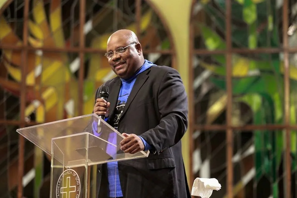 A man in a black blazer and blue shirt stands behind a clear podium with a church emblem, speaking into a microphone with a background of stained glass windows.