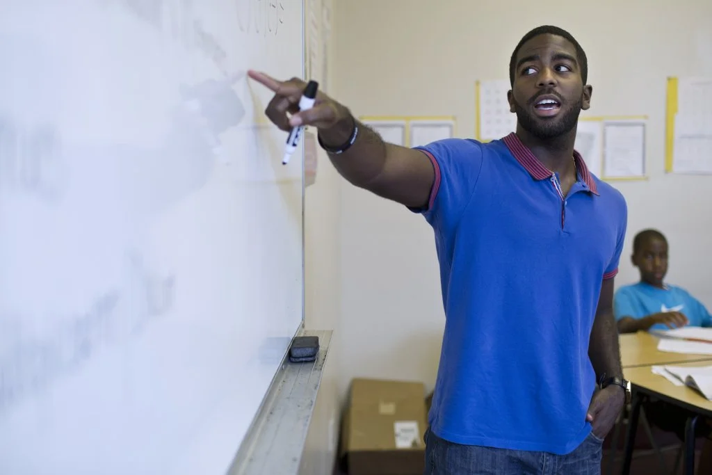 A male teacher pointing at a whiteboard during class with a marker in his hand, and a student sitting at a desk in the background.