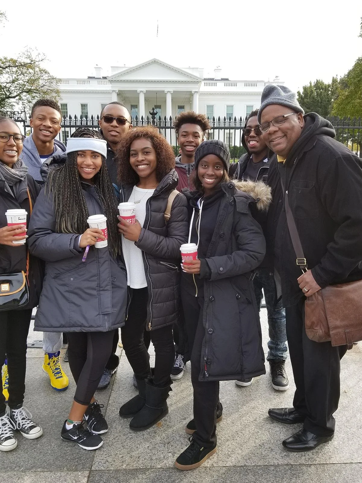 A group of diverse people, mostly young adults, standing in front of the White House, holding coffee cups and smiling for the camera.