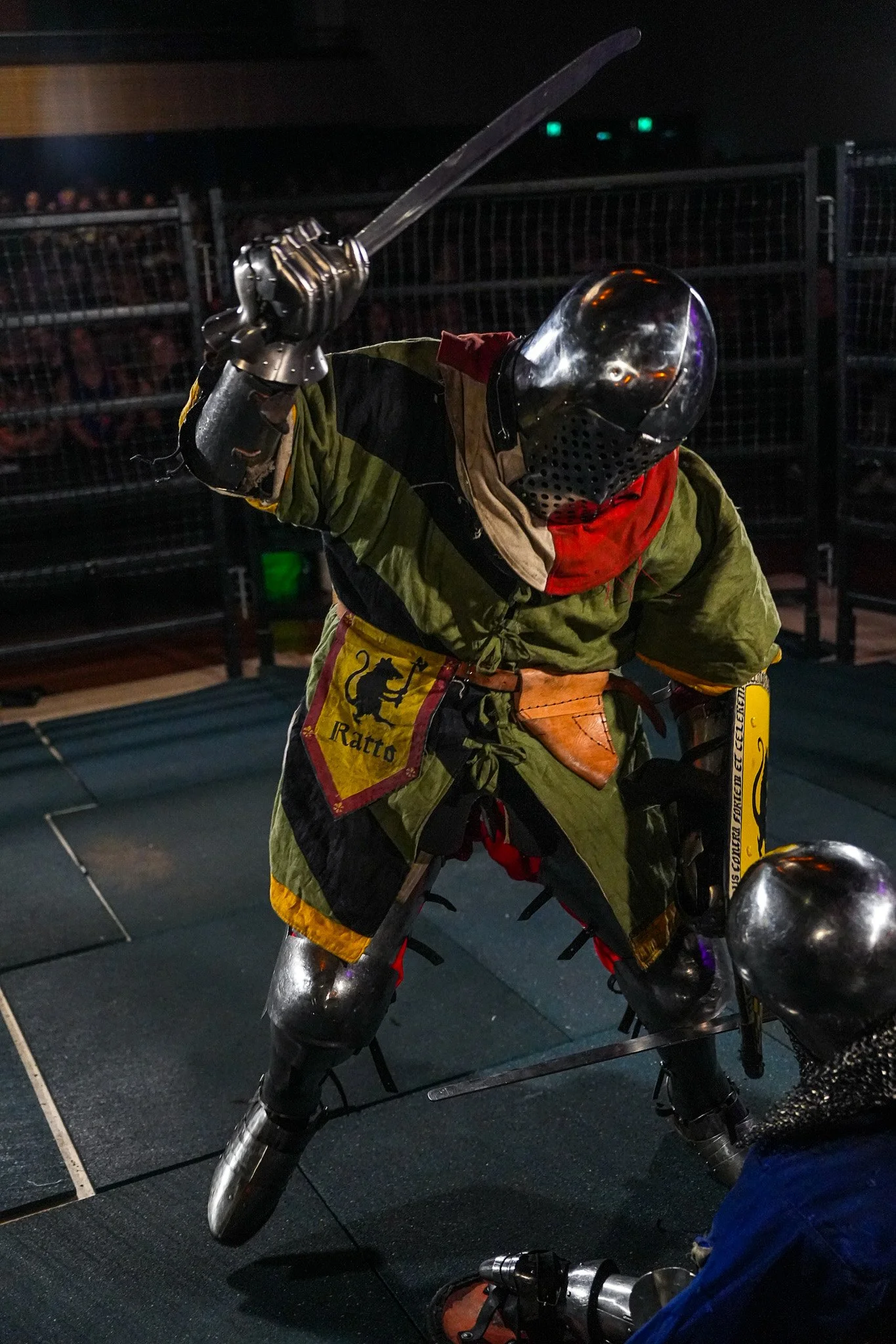 A person in a medieval knight costume with armor and a helmet, holding a sword, standing indoors with a crowd in the background.