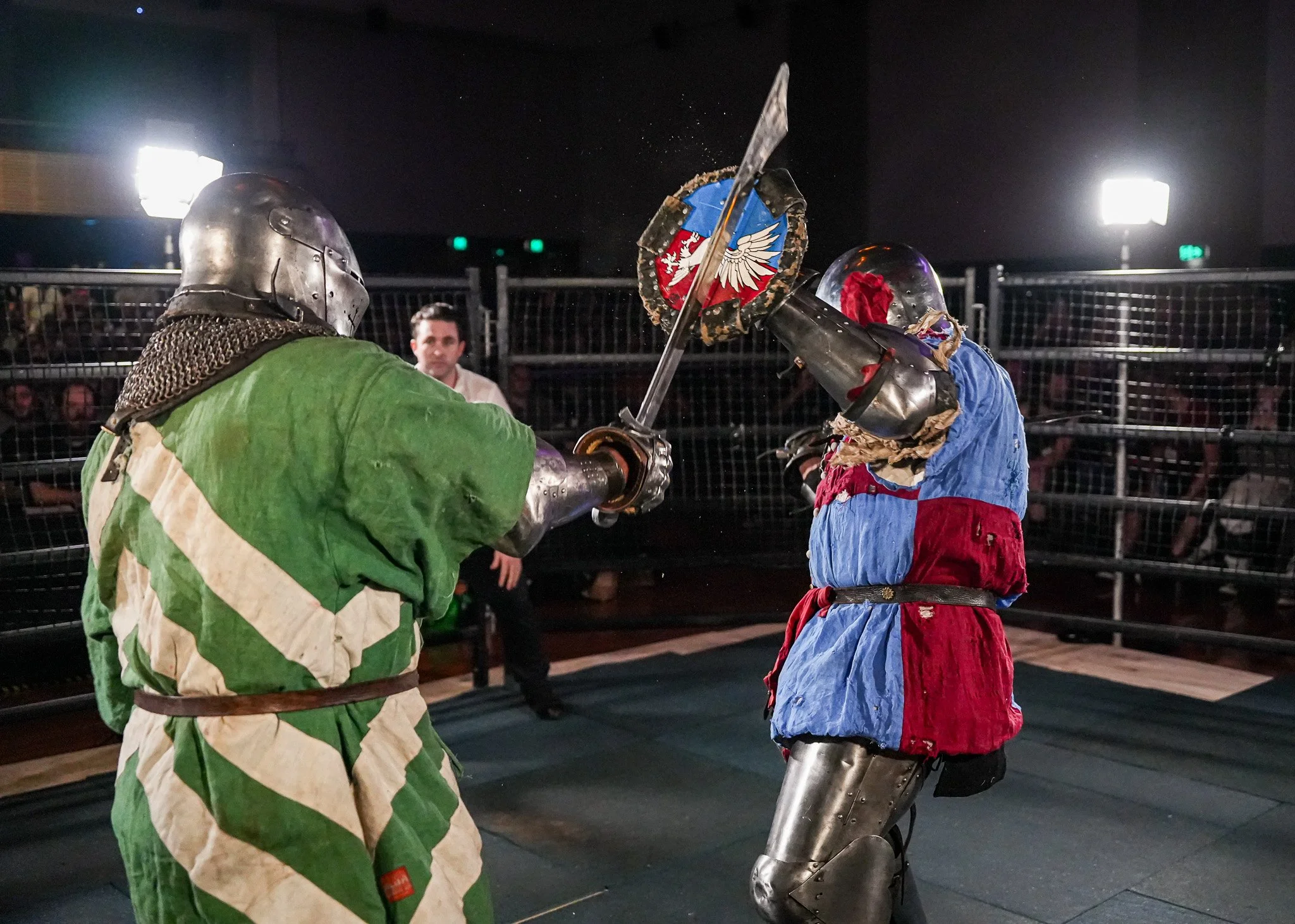 Two performers in knight costumes fighting with swords inside a ring, with an audience watching.