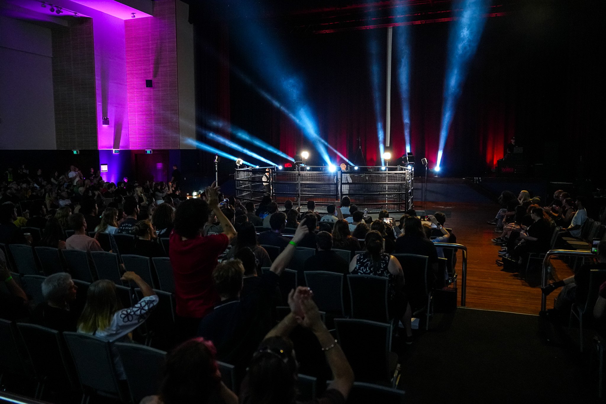 A wrestling ring inside an auditorium with spotlights and red curtains, crowd seated around watching the event.