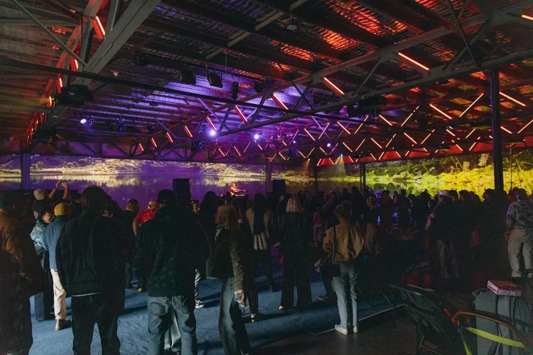 Crowd of people gathered at an indoor concert or event with colorful stage lighting and a large screen displaying a scenic landscape in the background.