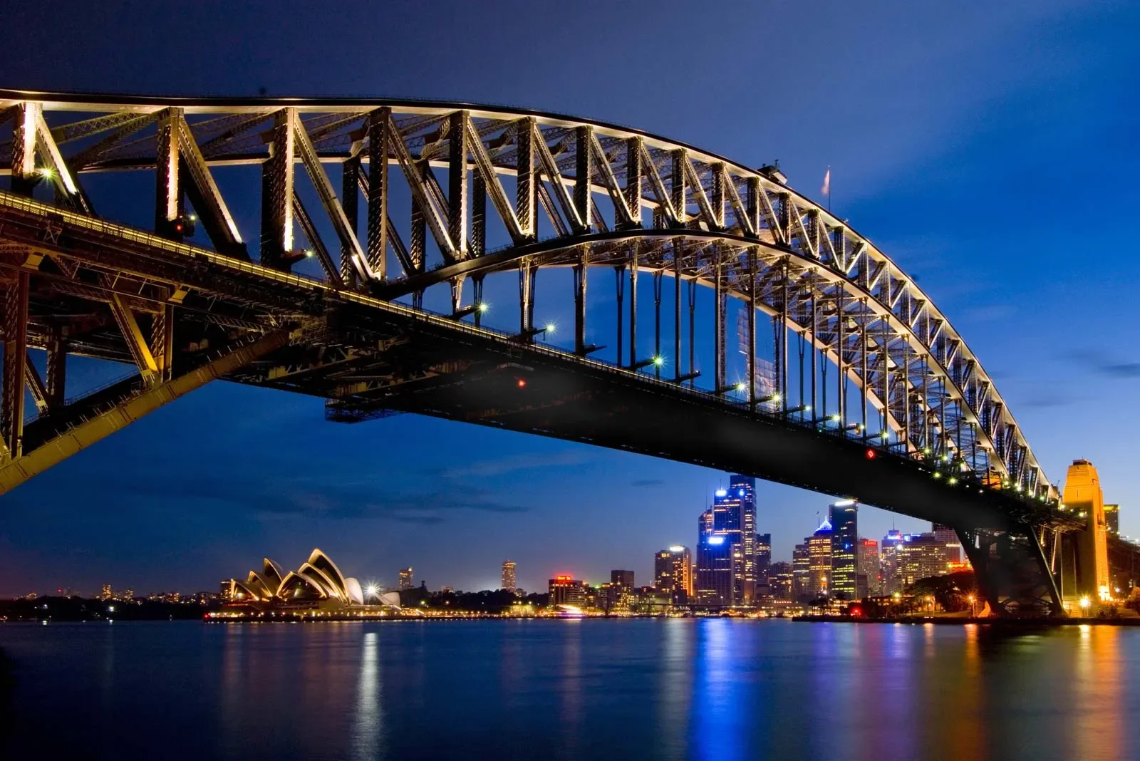 Night view of the Sydney Harbour Bridge with the city skyline and the Sydney Opera House reflected on the water.