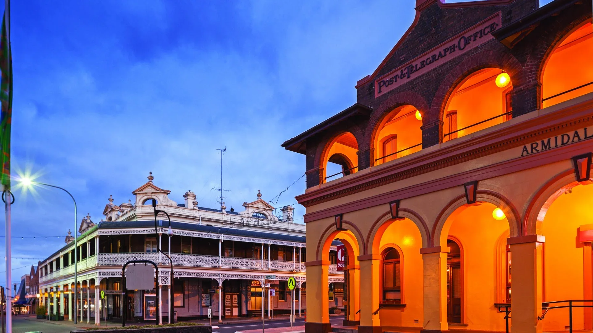 A historic building with arches illuminated in warm orange light, sign reading 'Post & Telegraph Office,' and a clock tower, against a twilight sky. Nearby structures feature Victorian architectural details.