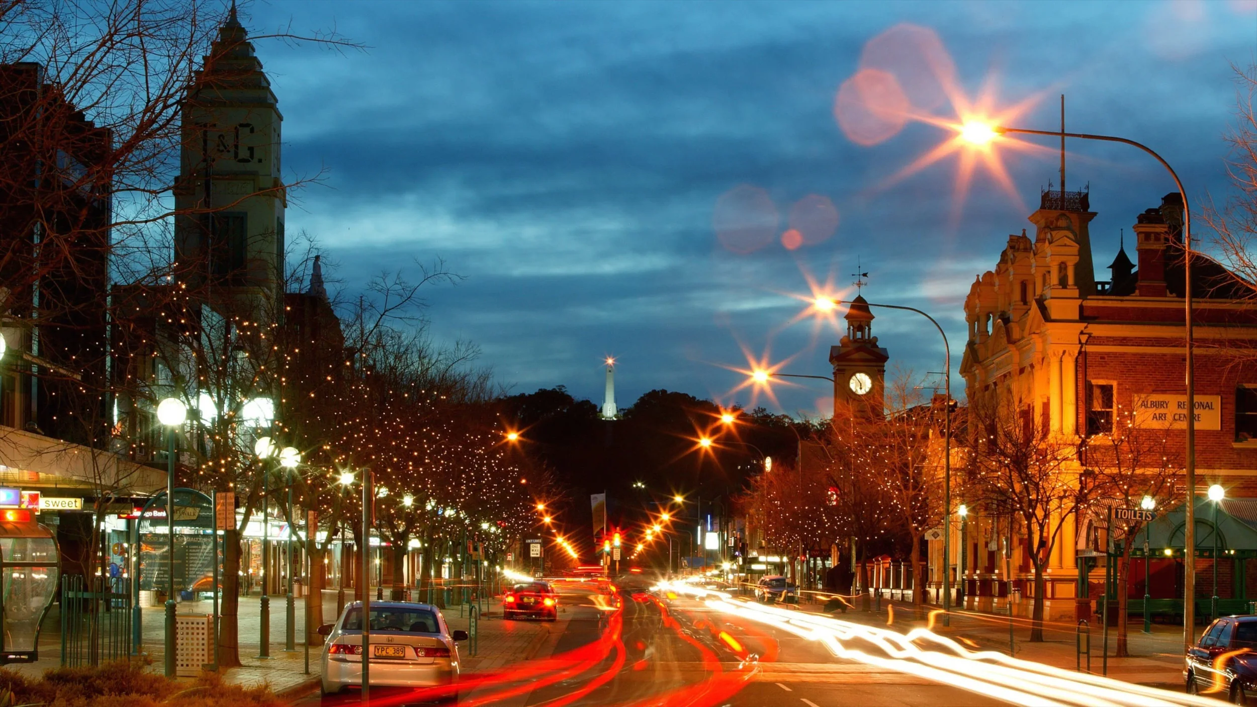 City street at sunset with illuminated streetlights, trees, historic buildings, and a prominent monument on the horizon.