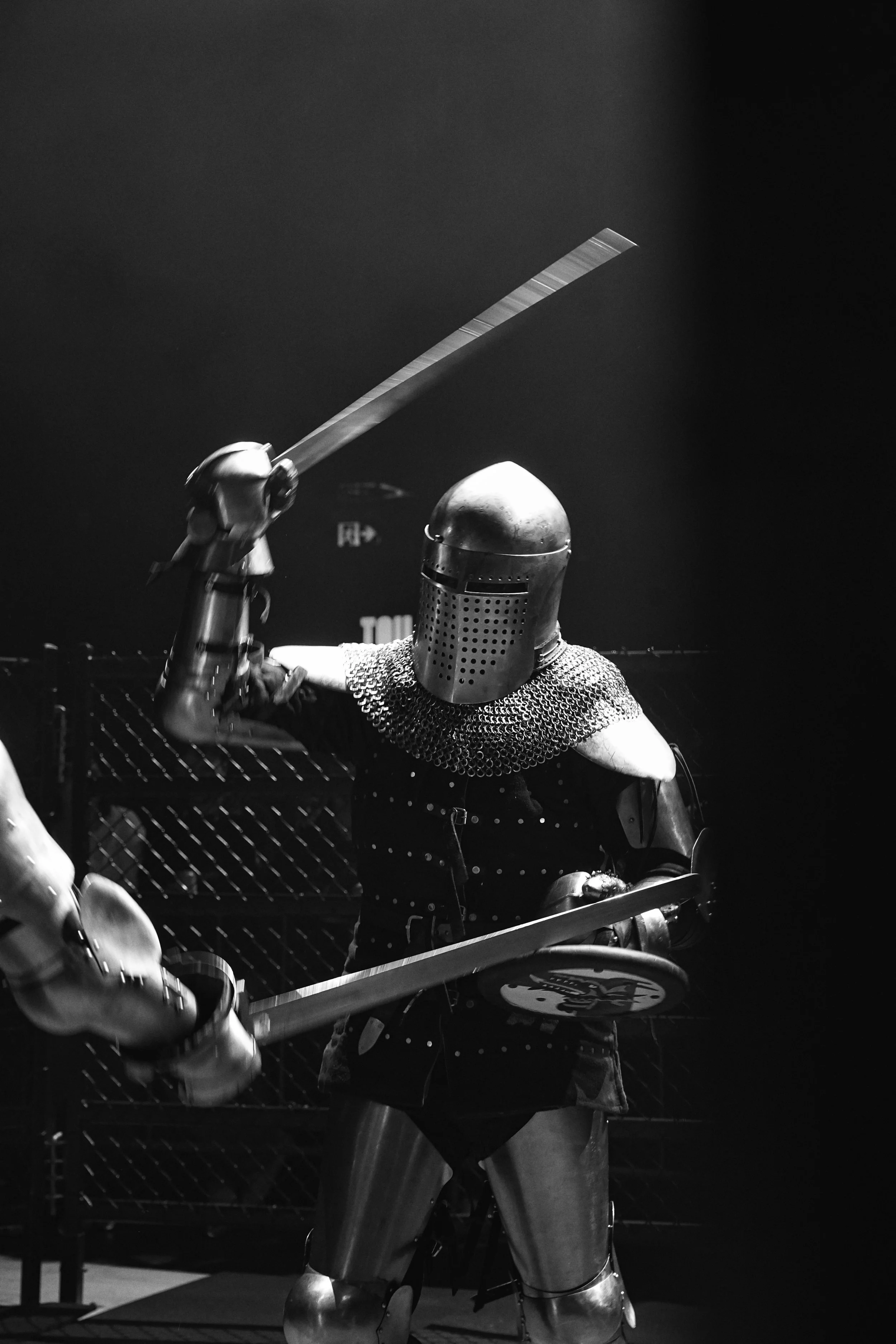 A person dressed in medieval knight armor, including a helmet and chainmail, holding a sword, standing behind a chainmail curtain, in a dark setting with a chain-link fence visible in the background.
