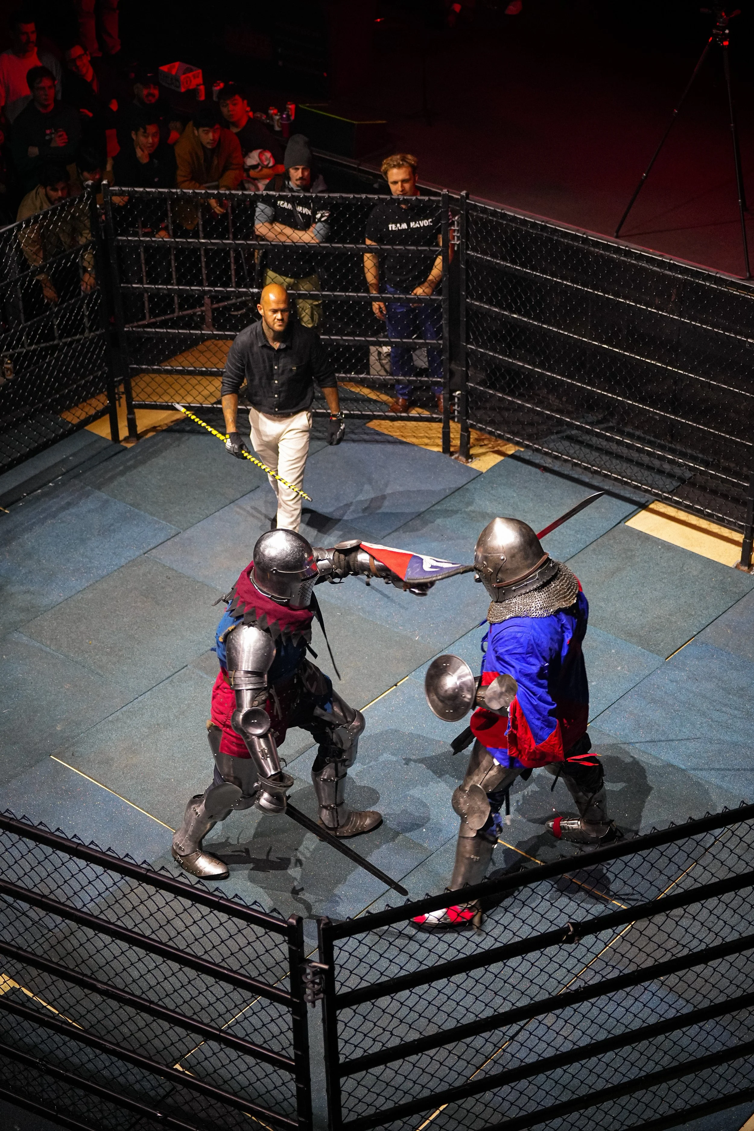 Two knights in armor engaging in a duel inside a fenced arena with a medieval theme. They are wearing helmets and medieval-style clothing, while onlookers observe from outside the fencing.