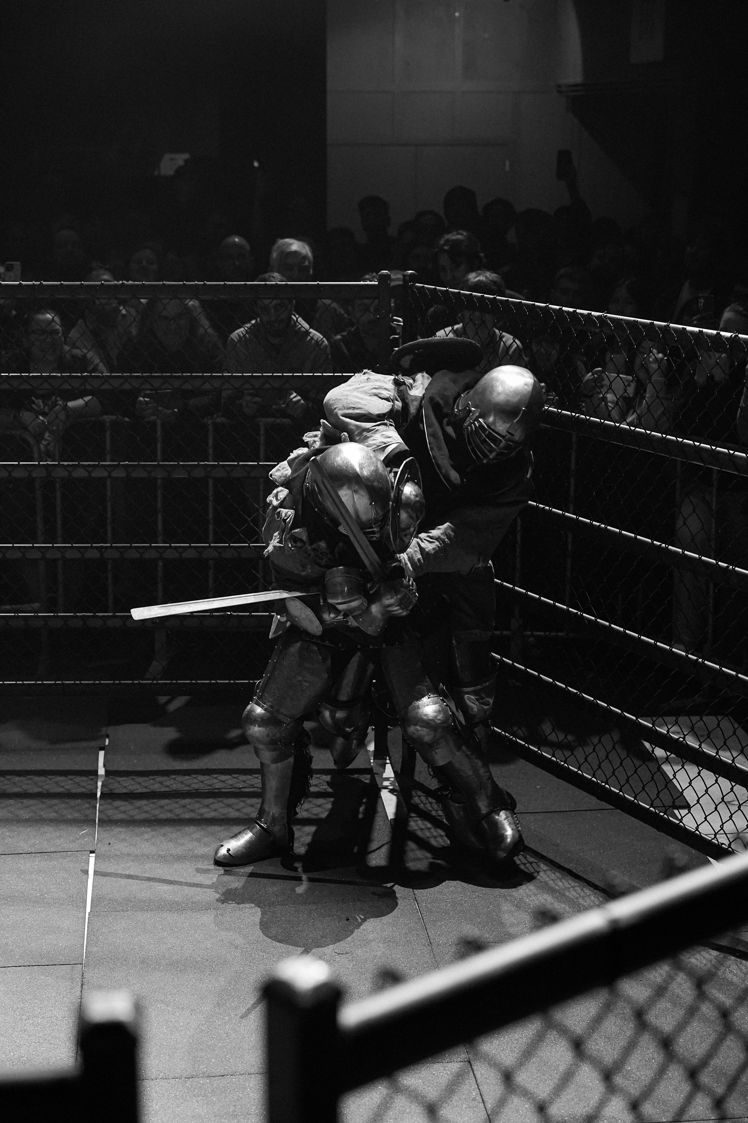 A firefighter in full protective gear, including a helmet, working with a chainsaw near a fence fence at night. Behind the fence, a crowd of people is gathered, with some visible faces and one person holding an object in the air. The scene is dark, with limited lighting.