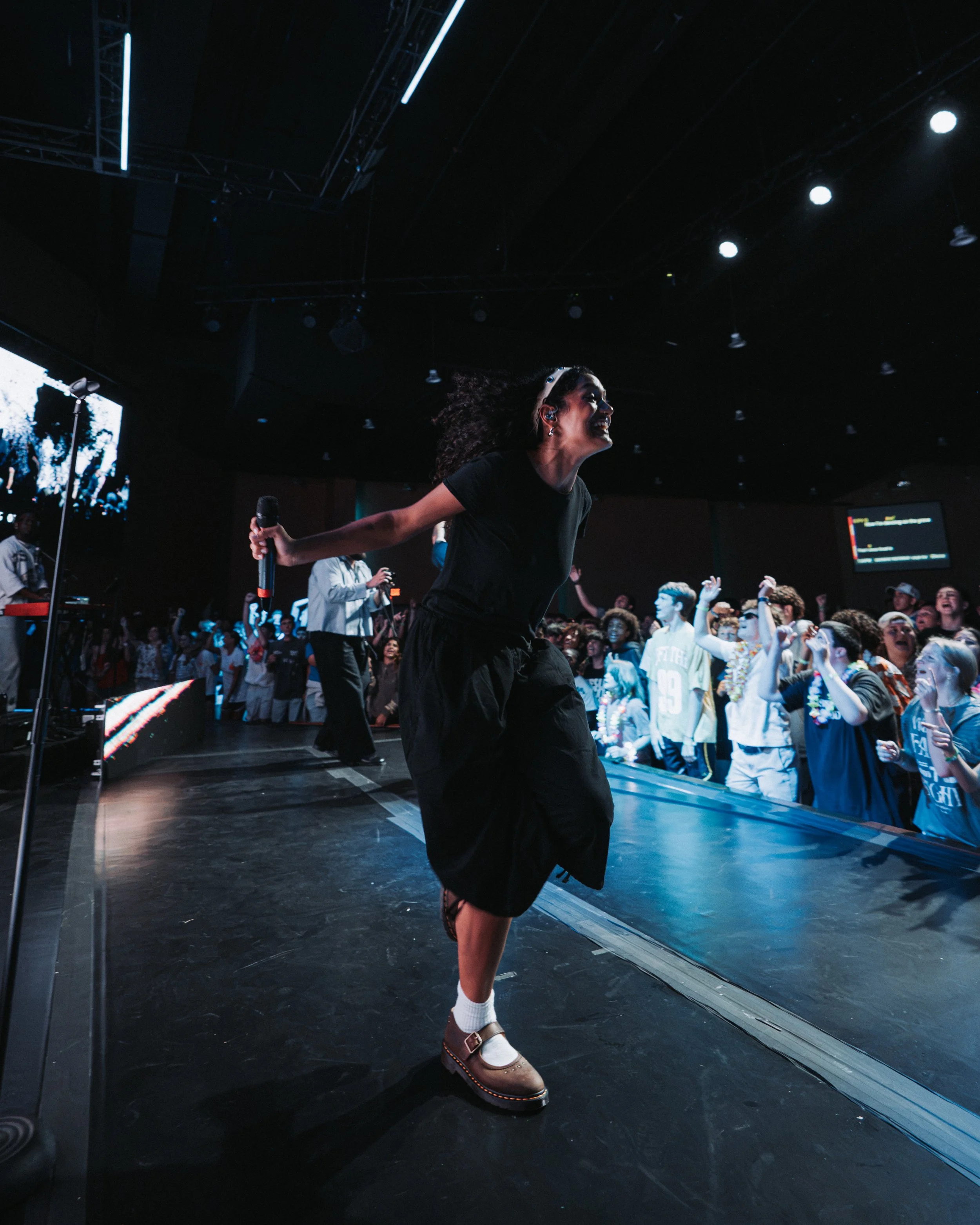 A woman in a black dress and white socks with brown shoes is dancing energetically on a stage at a concert or event, with an audience behind her. She is holding a microphone in her right hand, smiling, with her curly hair flowing. The background shows a large screen and bright stage lights in a dark venue.